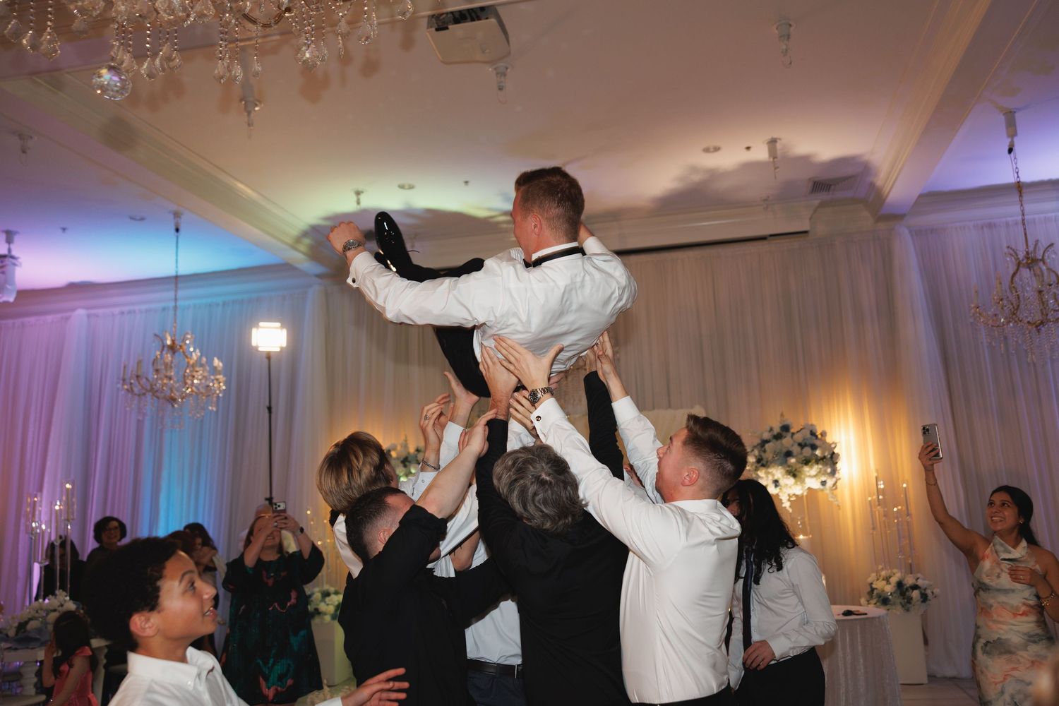 Groomsmen lift the groom into the air during a lively wedding reception celebration in a decorated ballroom at The Yorkmont in Charlotte, NC
