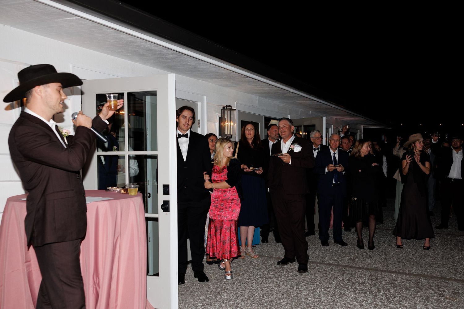 Groom in black cowboy hat raises champagne glass during wedding toast while guests celebrate outdoors at night.