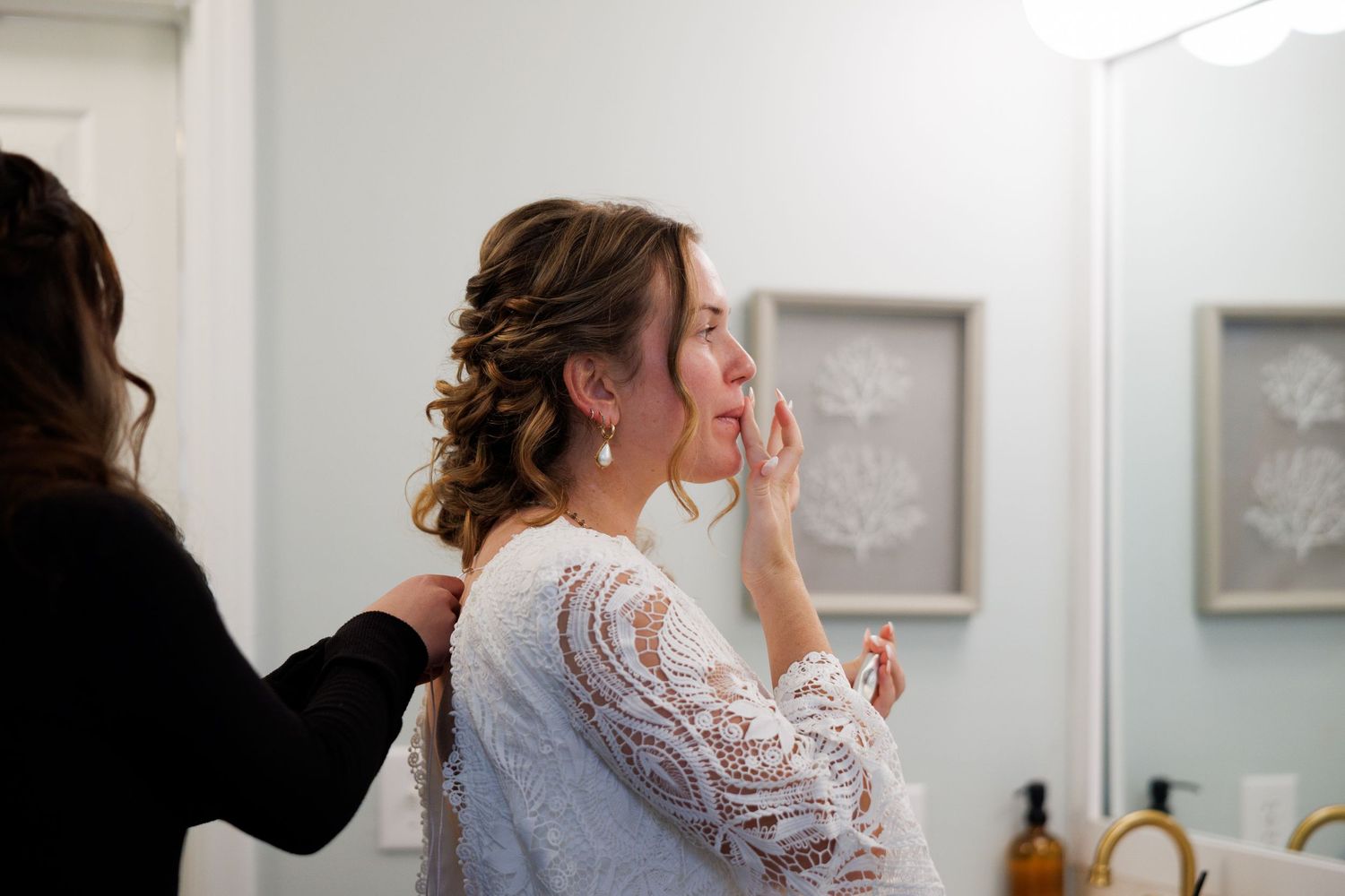 Bride getting ready as bridesmaid helps with her lace wedding dress in a bright bathroom mirror.