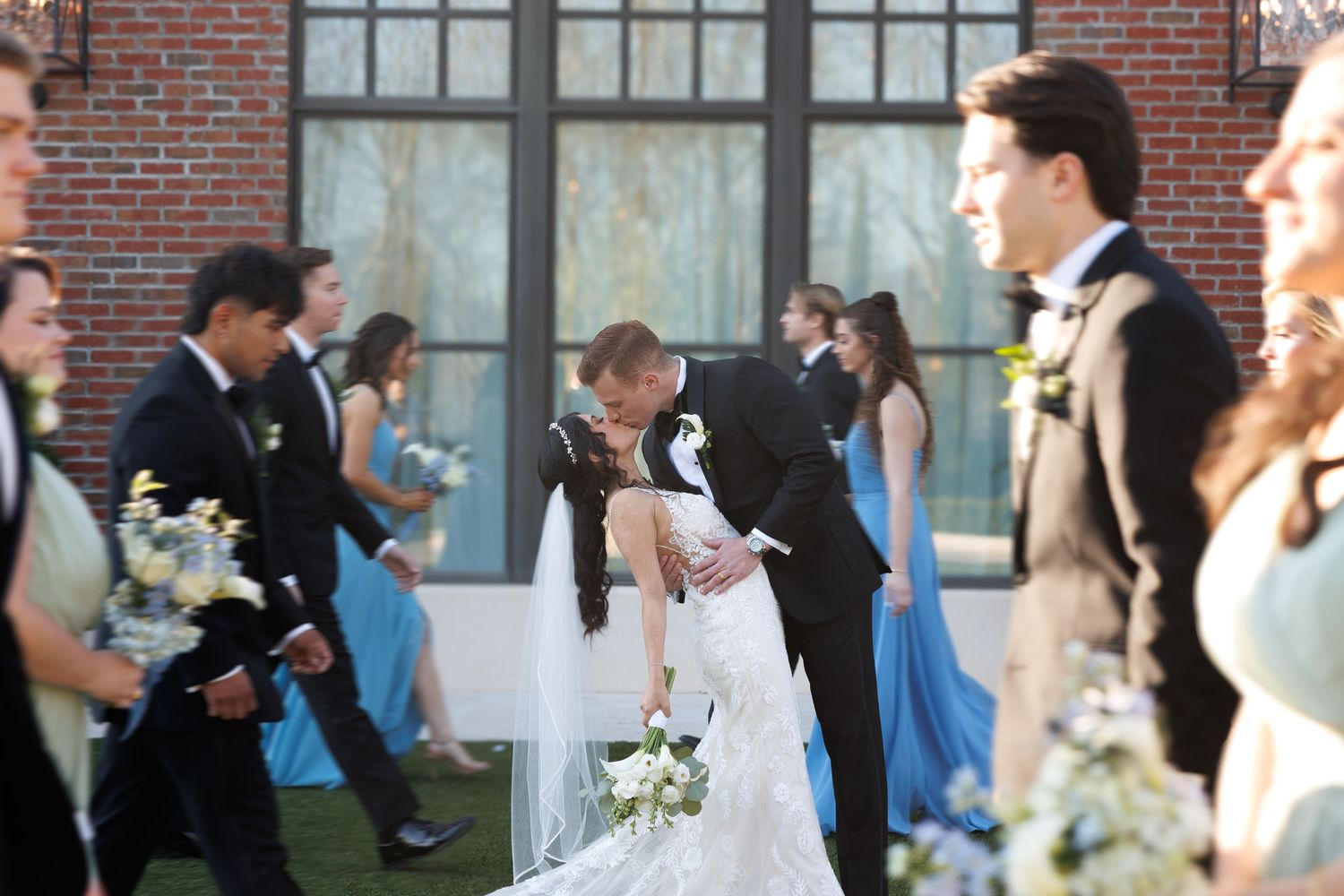 Bride and groom share a romantic kiss surrounded by wedding party in blue dresses outside the brick courtyard at the Yorkmont in Charlotte.