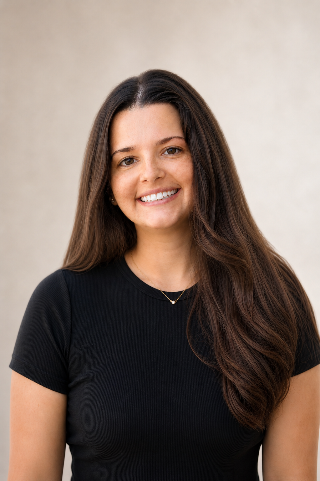 Smiling young woman with long brown hair wearing a black t-shirt and gold necklace against a neutral background.