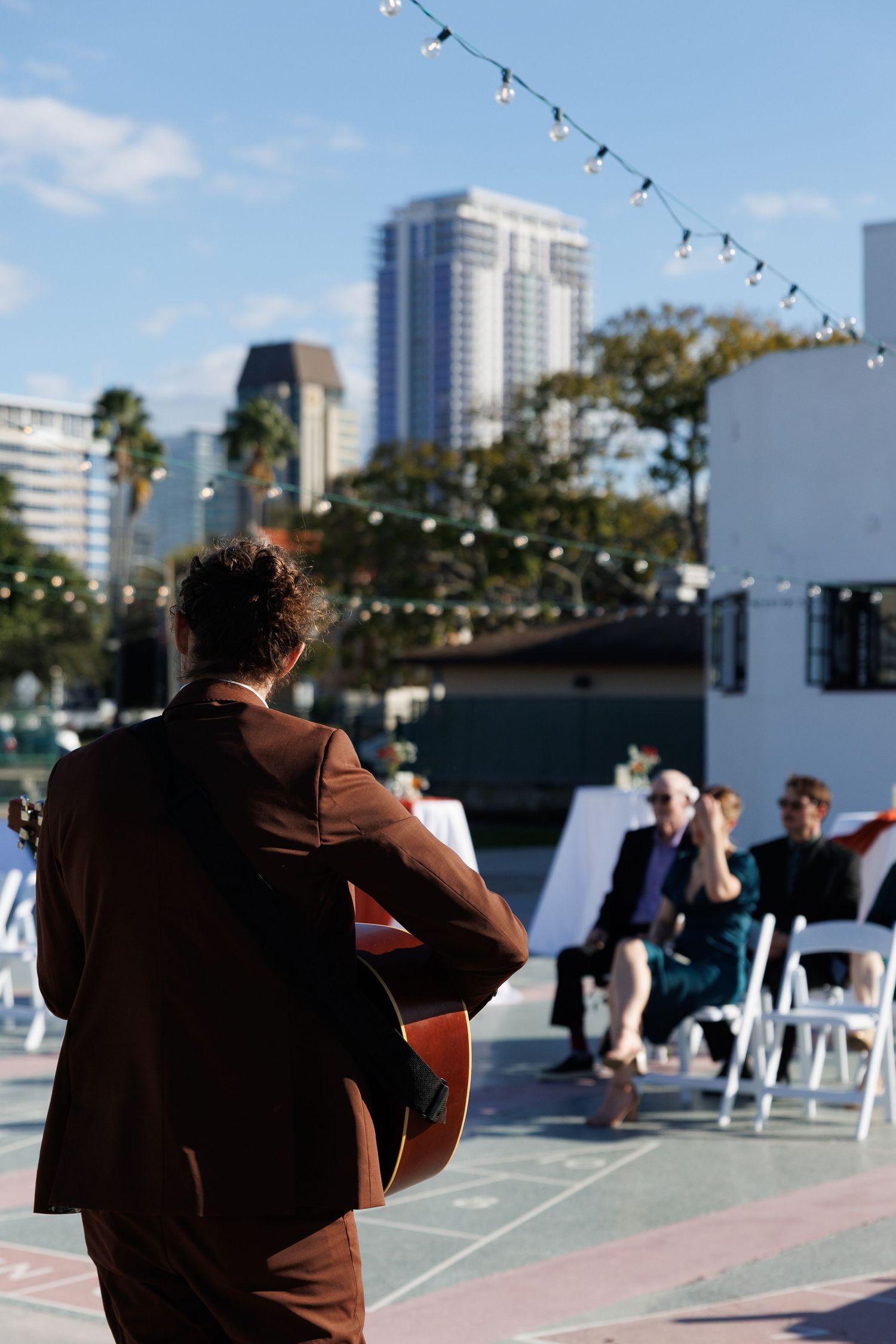 Guitarist in brown suit performs at outdoor wedding ceremony with city skyline backdrop.