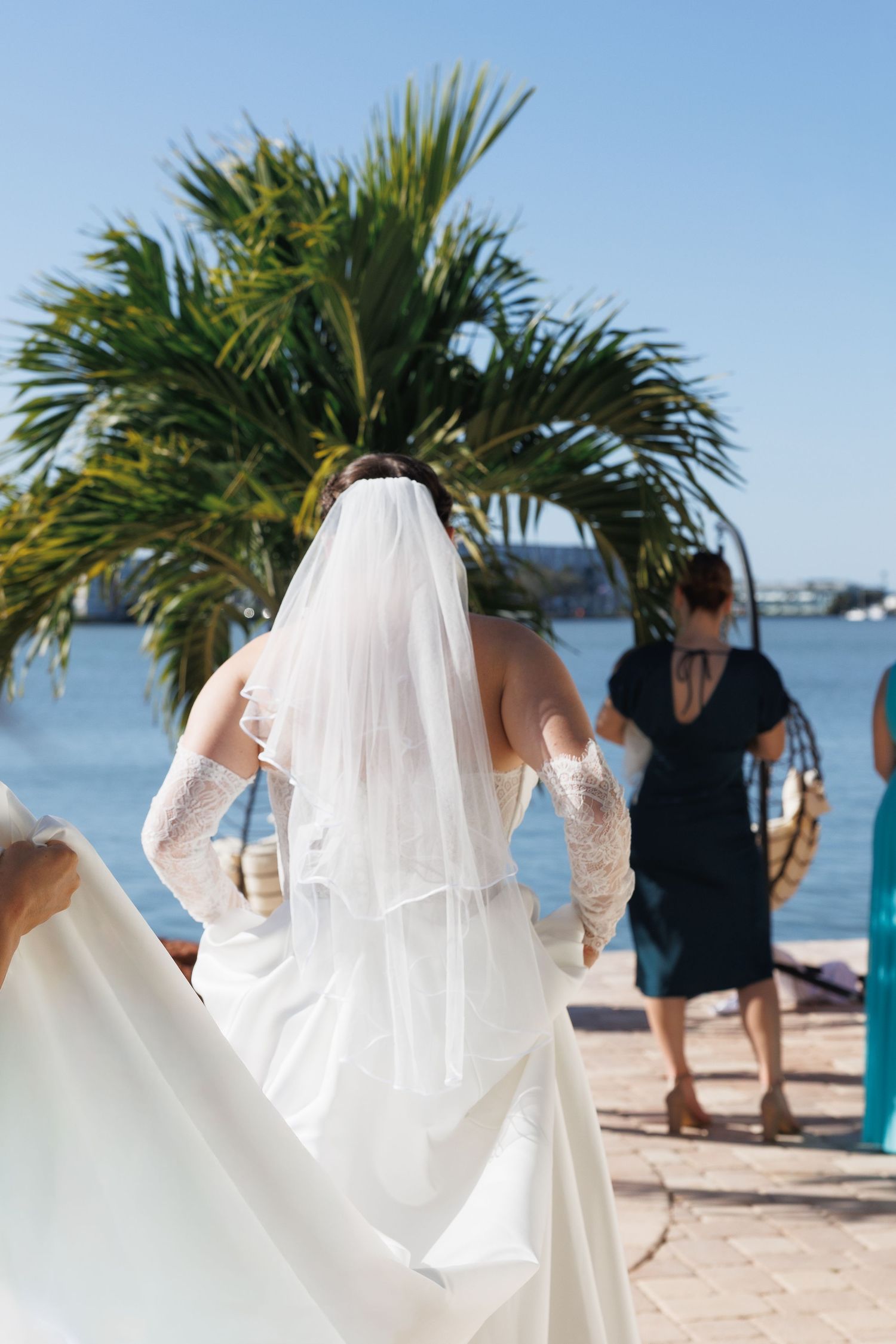 Bride in white gown and veil walks toward waterfront palm trees at outdoor wedding ceremony.