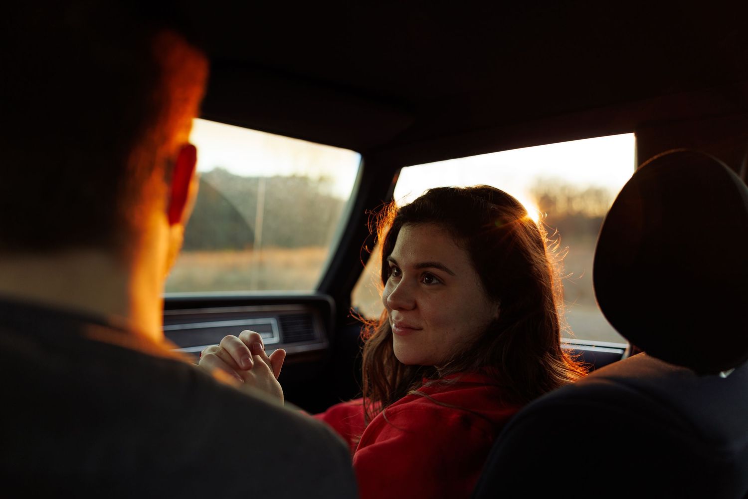 Young woman with red hair smiling warmly at a man inside a car during golden hour.