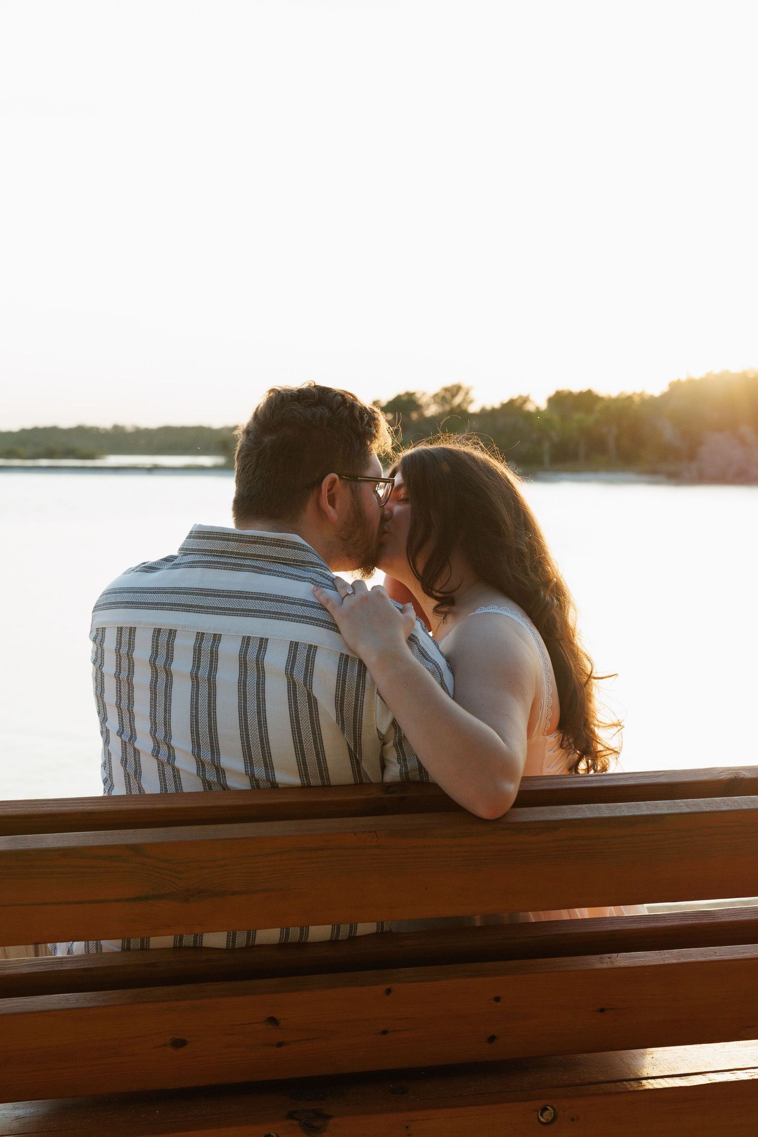 Couple kissing on a wooden bench at sunset by a serene lake with warm golden light.