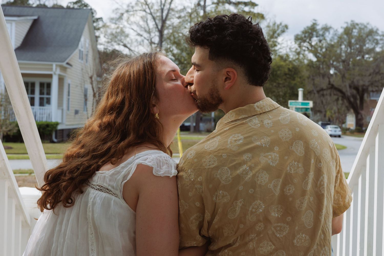 A couple shares a romantic kiss on a white porch railing in a charming residential neighborhood.
