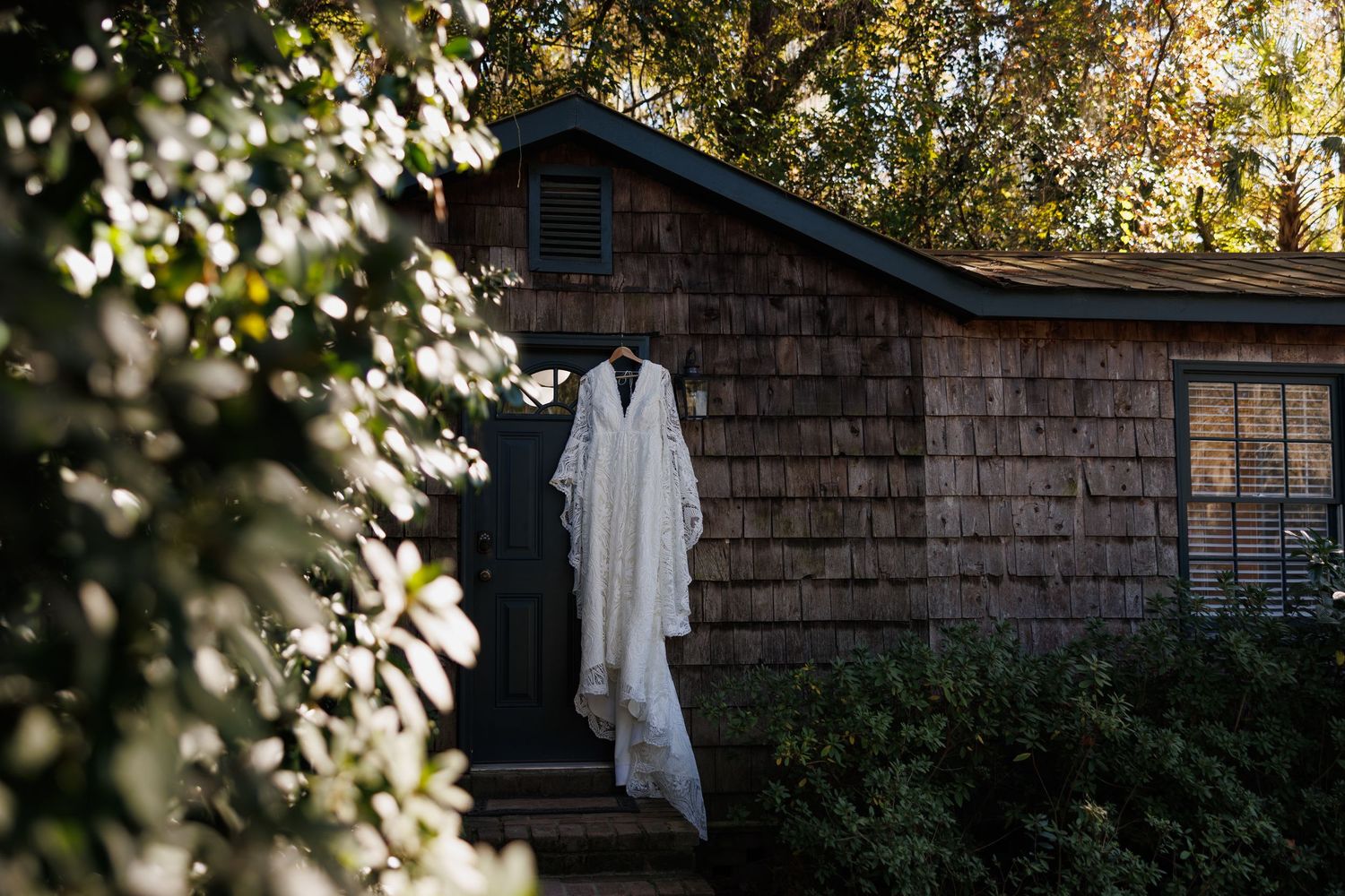White wedding gown hanging on rustic cedar shake cottage door surrounded by lush green trees in soft natural light.