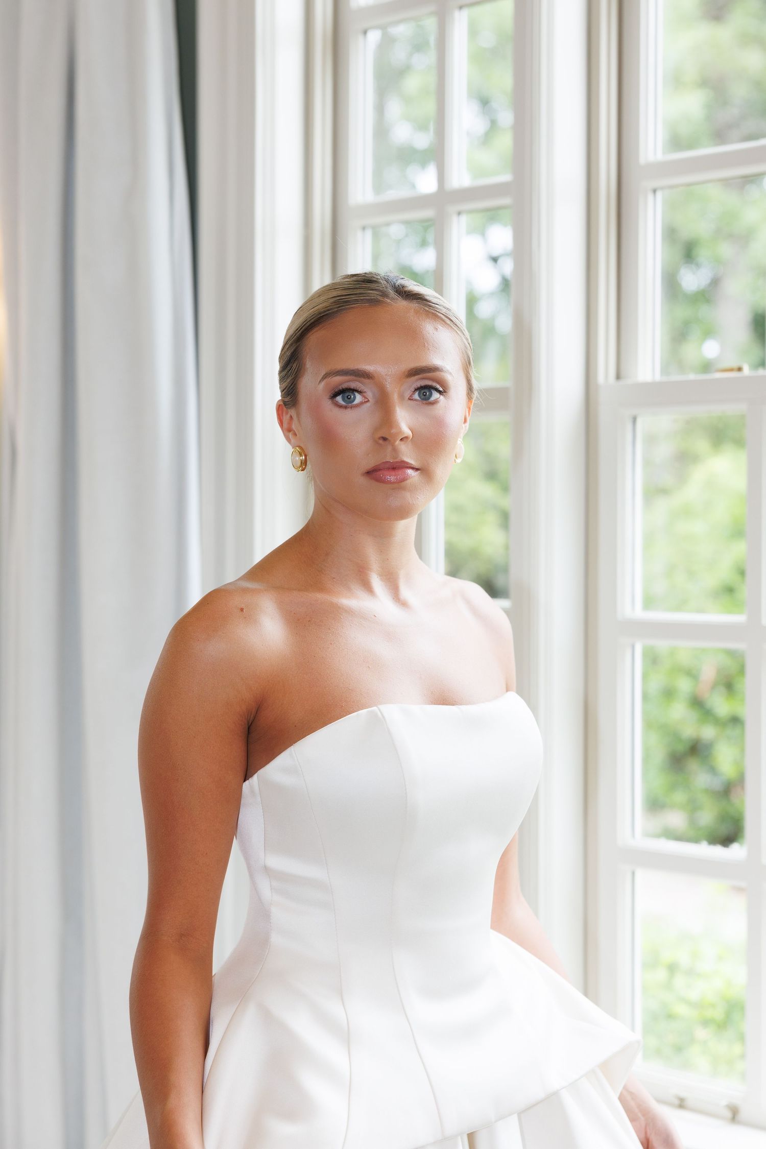 Elegant bride in strapless white wedding gown with gold earrings poses by bright window with soft natural light.