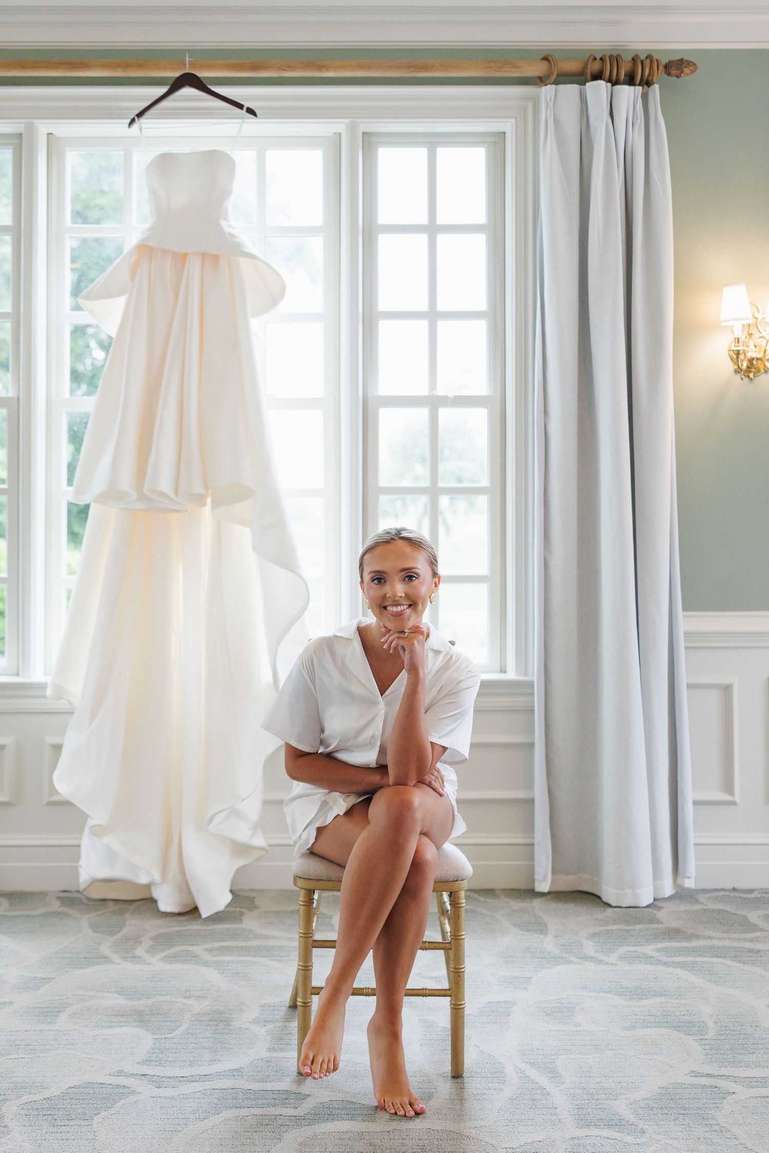 Smiling bride in white robe sits barefoot on chair before large window with elegant strapless wedding gown hanging behind her.
