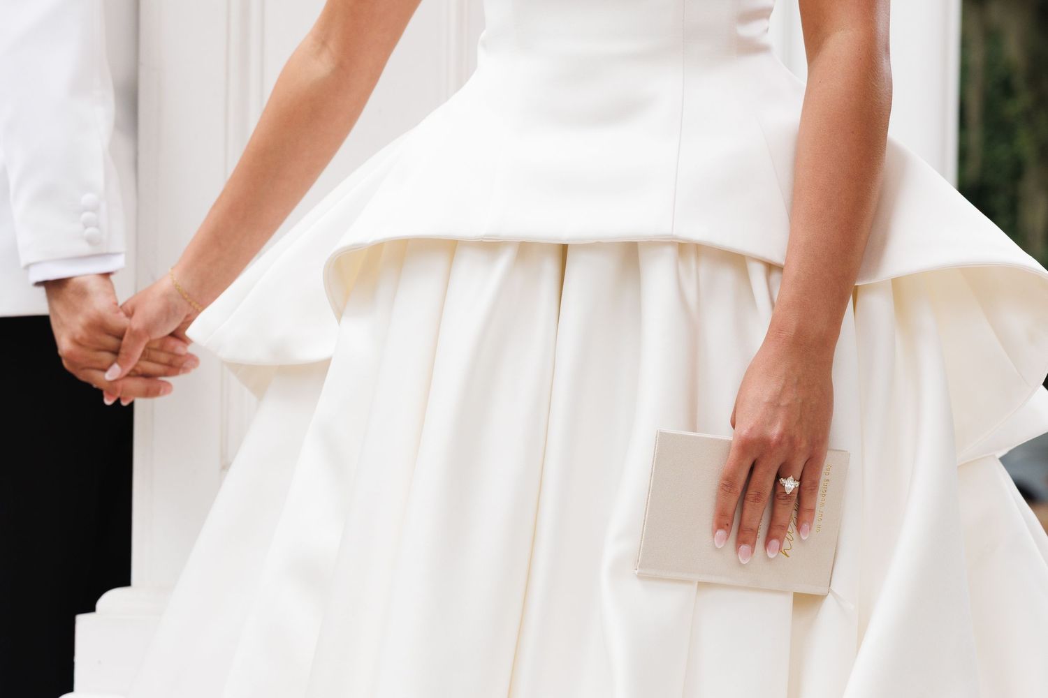 Bride in white wedding gown holding a clutch purse and groom's hand, featuring an elegant tiered skirt design.