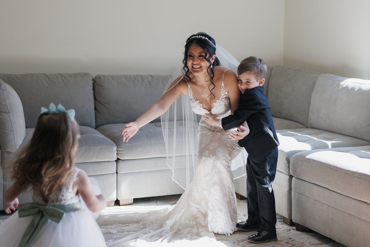 Bride in a lace wedding gown and veil laughing with a flower girl and ring bearer in a sunlit room.