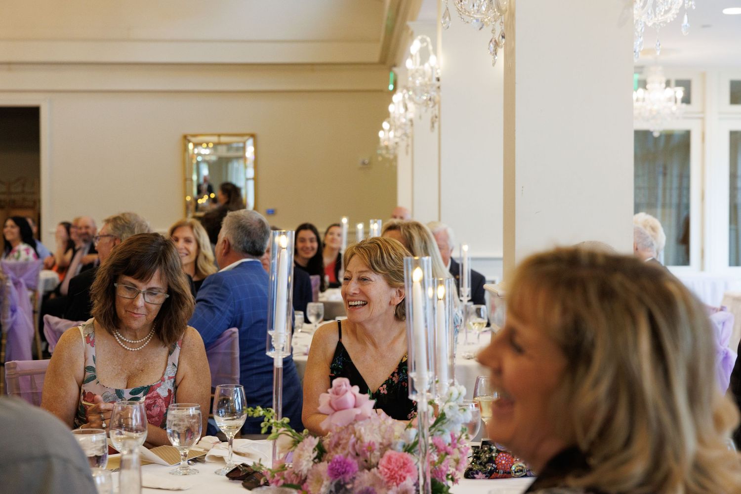 Women laughing and socializing at an elegant formal dinner event with floral centerpieces and chandeliers.