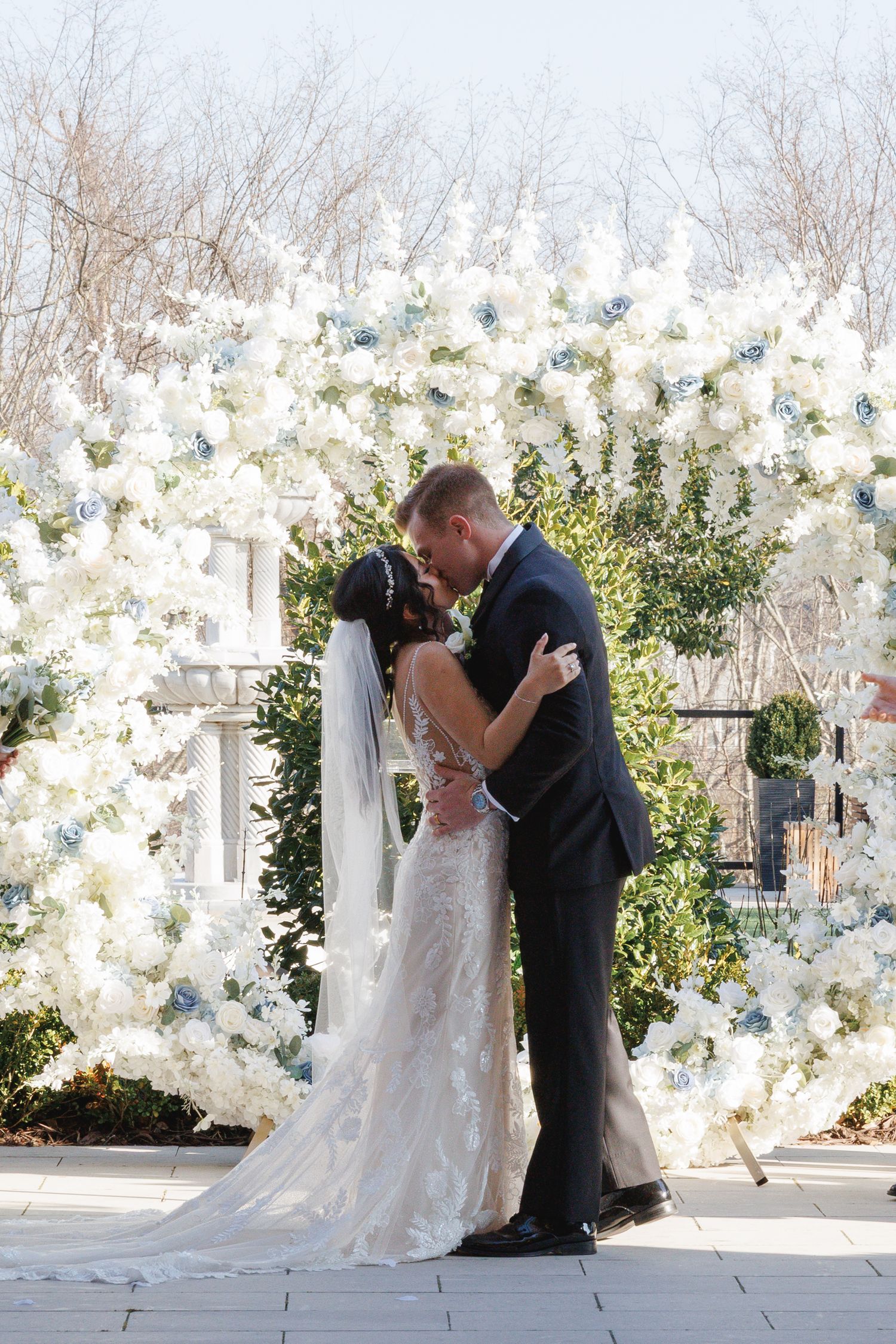 Bride and groom sharing first kiss beneath stunning white and blue floral wedding arch outdoors at The Yorkmont in Charlotte, NC.