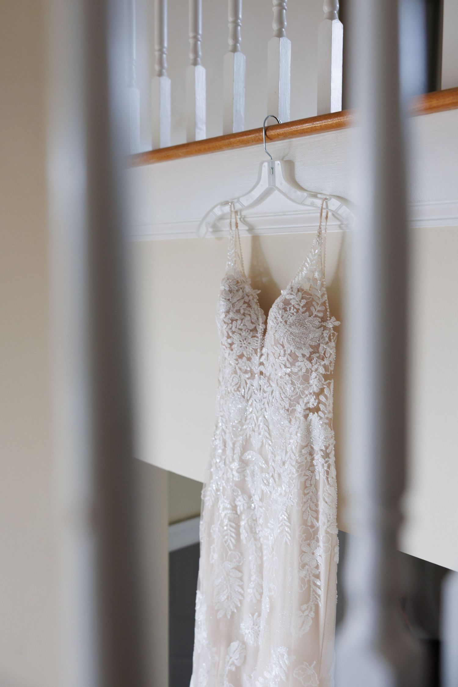 Delicate lace and beaded wedding gown hanging on a clear hanger over a staircase railing indoors.