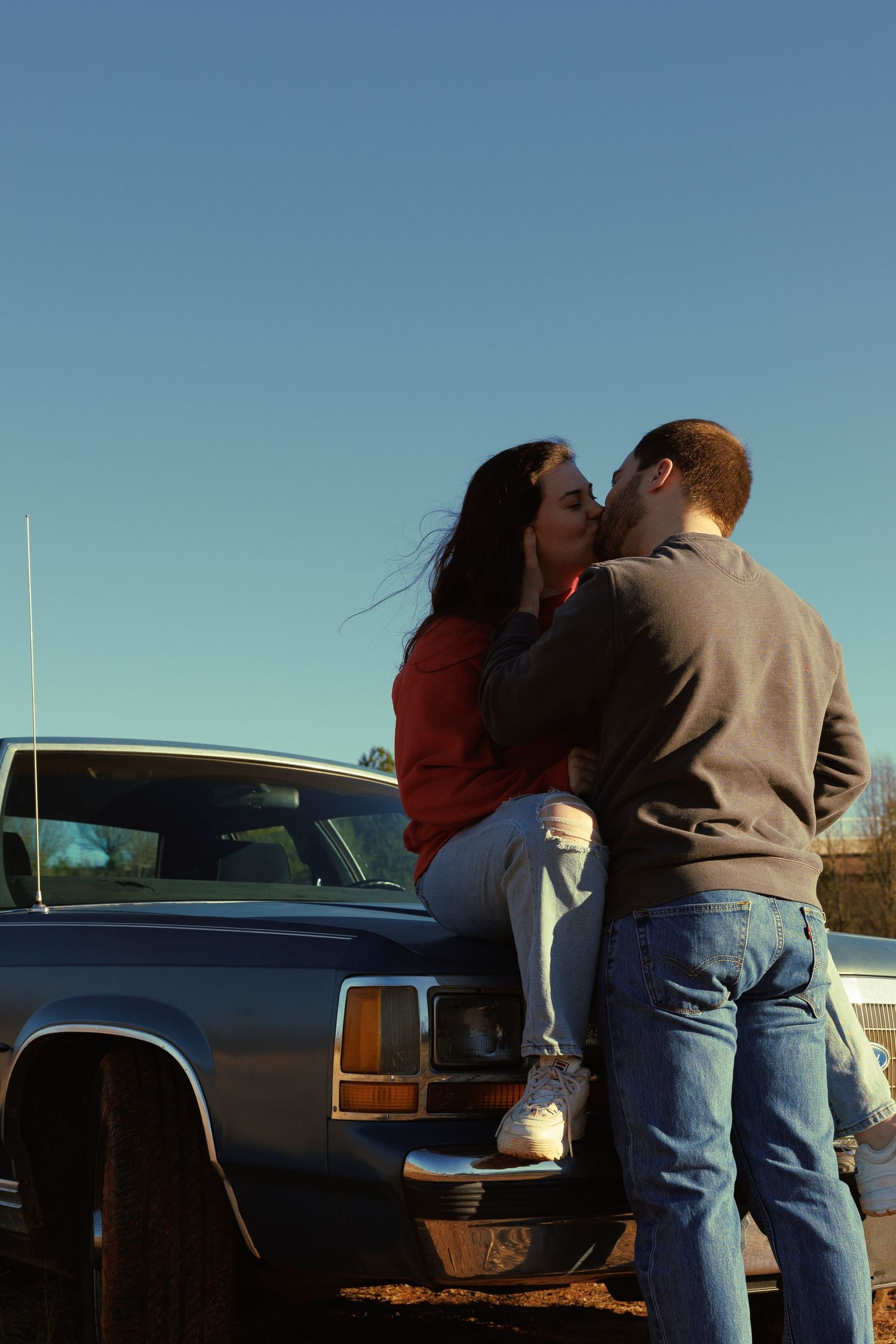 A couple sharing a romantic kiss beside a vintage blue car outdoors at golden hour.