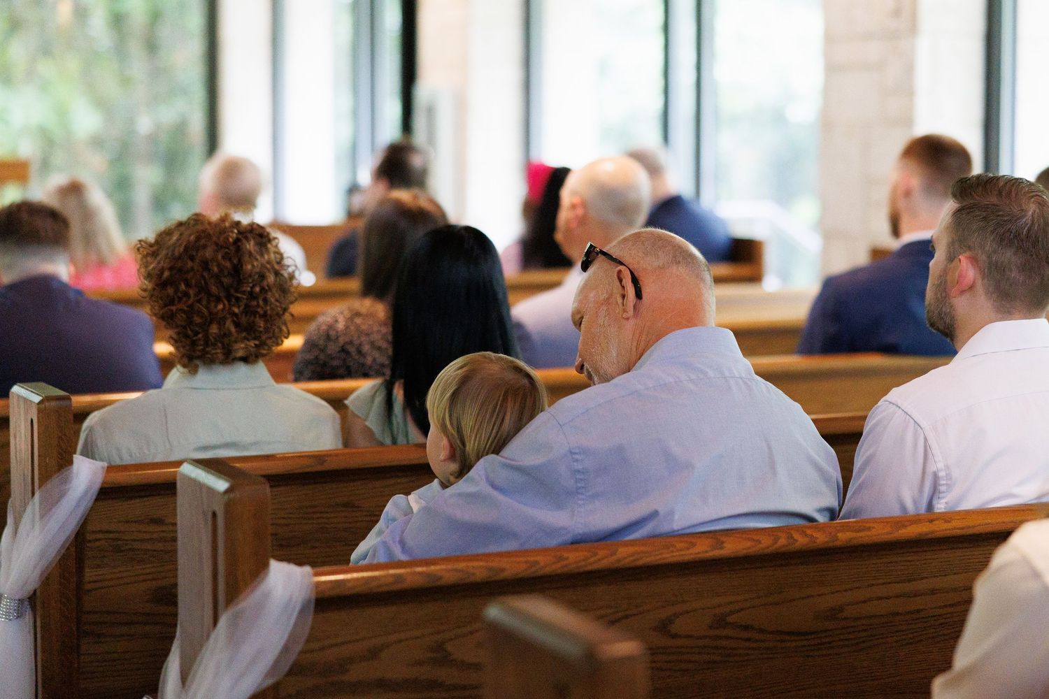 Congregation seated in wooden church pews during a religious service, with an elderly couple embracing in the foreground.