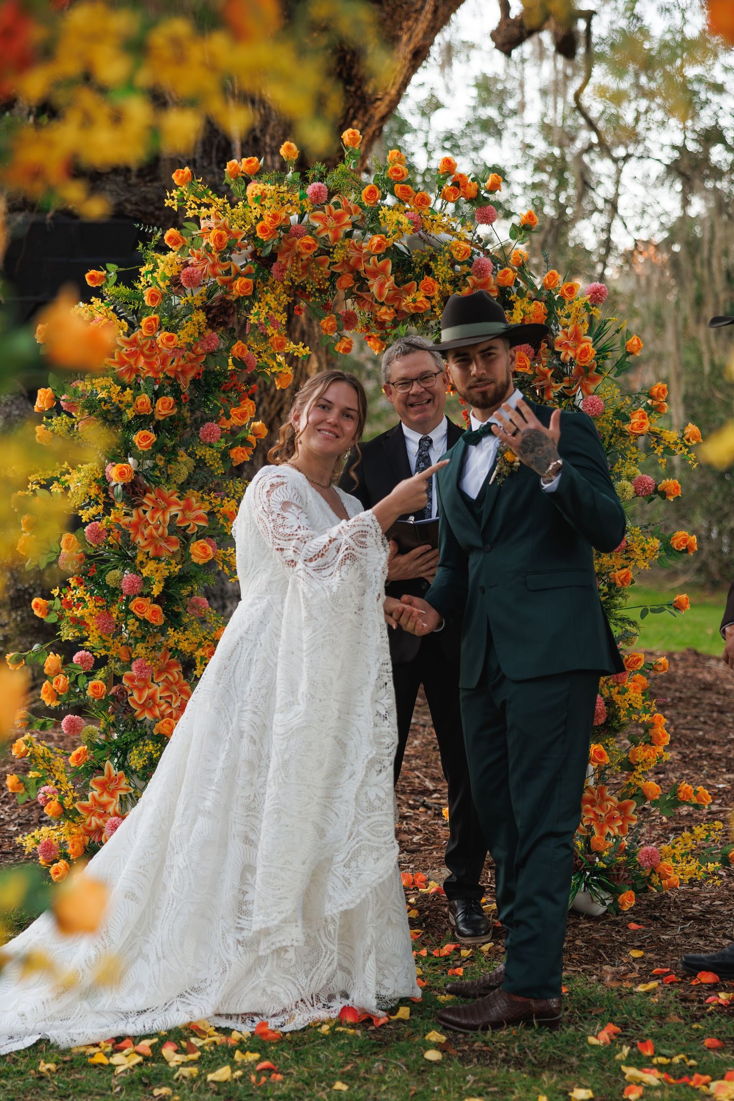 Bride and groom posing with officiant under vibrant orange floral arch during outdoor fall wedding ceremony.