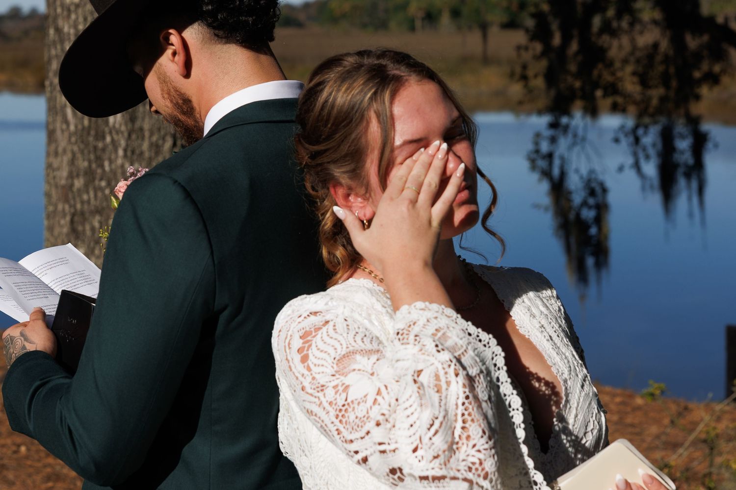Emotional bride in lace wedding dress wiping tears during outdoor lakeside wedding ceremony.