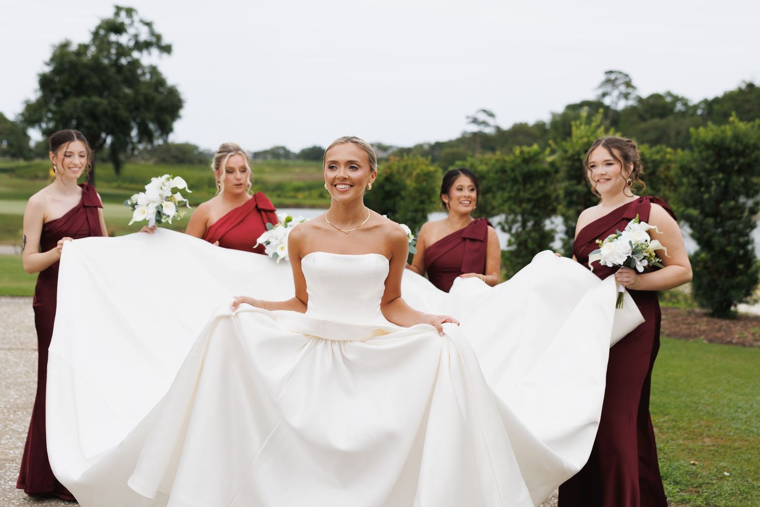 Smiling bride in strapless gown surrounded by bridesmaids in burgundy dresses spreading her long train outdoors.