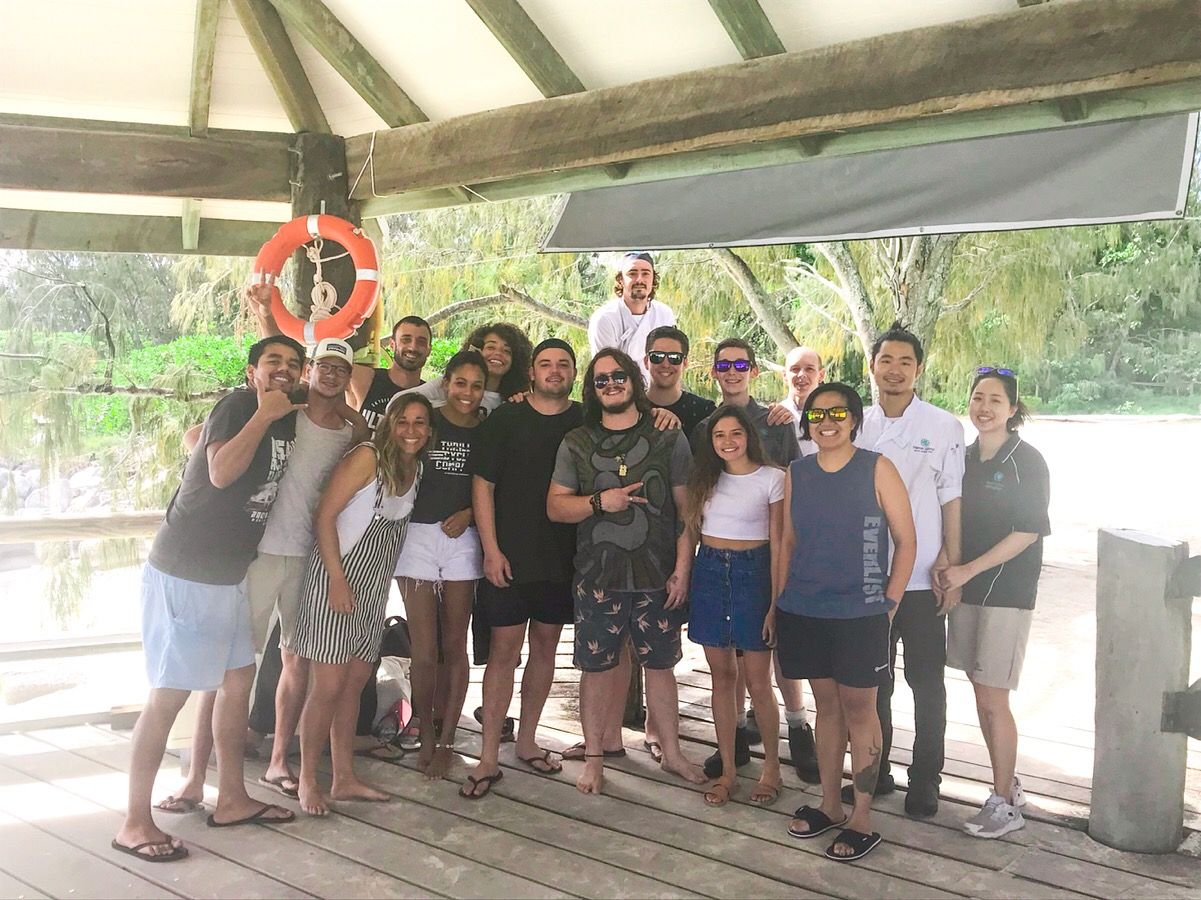 Large group of young adults posing together on a tropical waterfront pavilion with lush greenery in the background.