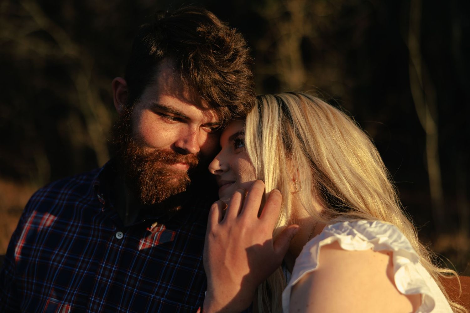 Bearded man in plaid shirt tenderly touches blonde woman's face during golden hour outdoor portrait.
