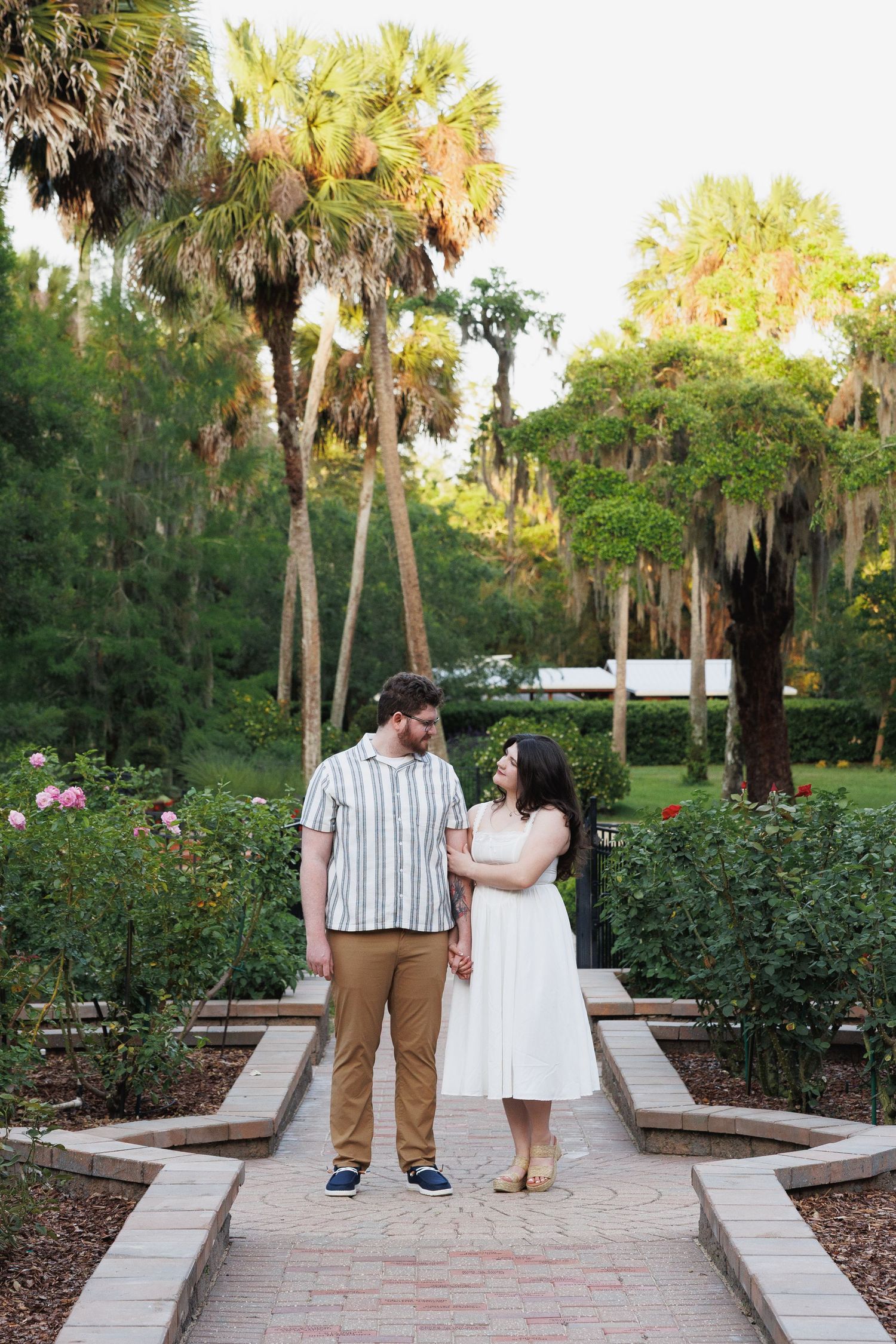 Couple holding hands in a lush garden with palm trees, man in striped shirt and woman in white dress.