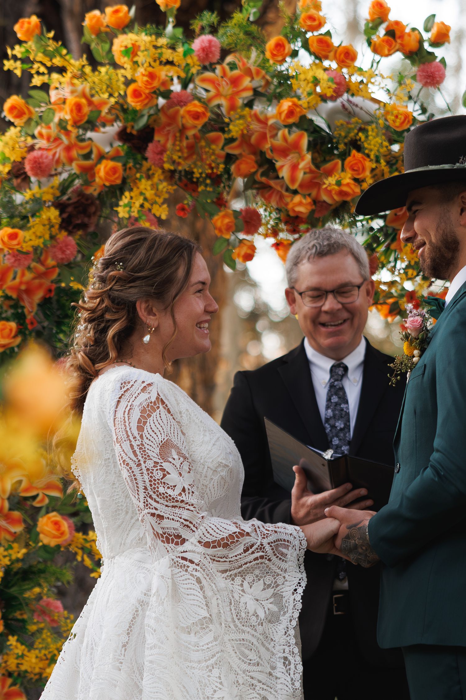 Bride and groom exchanging rings at outdoor ceremony beneath vibrant orange and yellow floral arch.