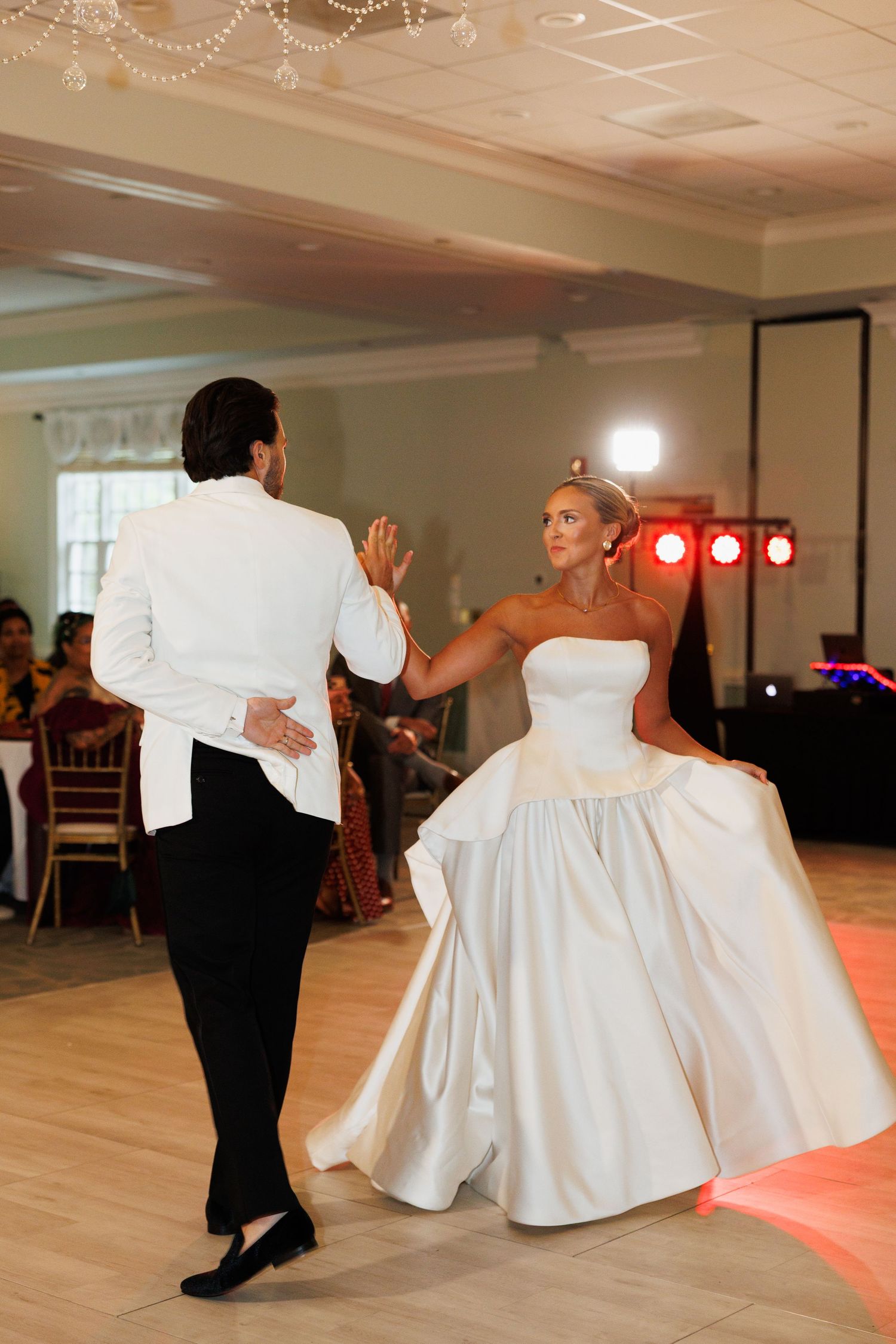 Bride and groom share their first dance at wedding reception, bride in strapless ball gown, groom in white jacket.