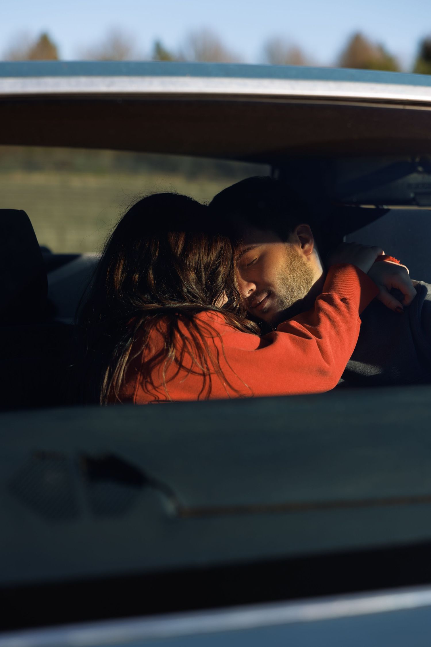 A couple shares an intimate embrace inside a car, bathed in warm golden sunlight through the window.