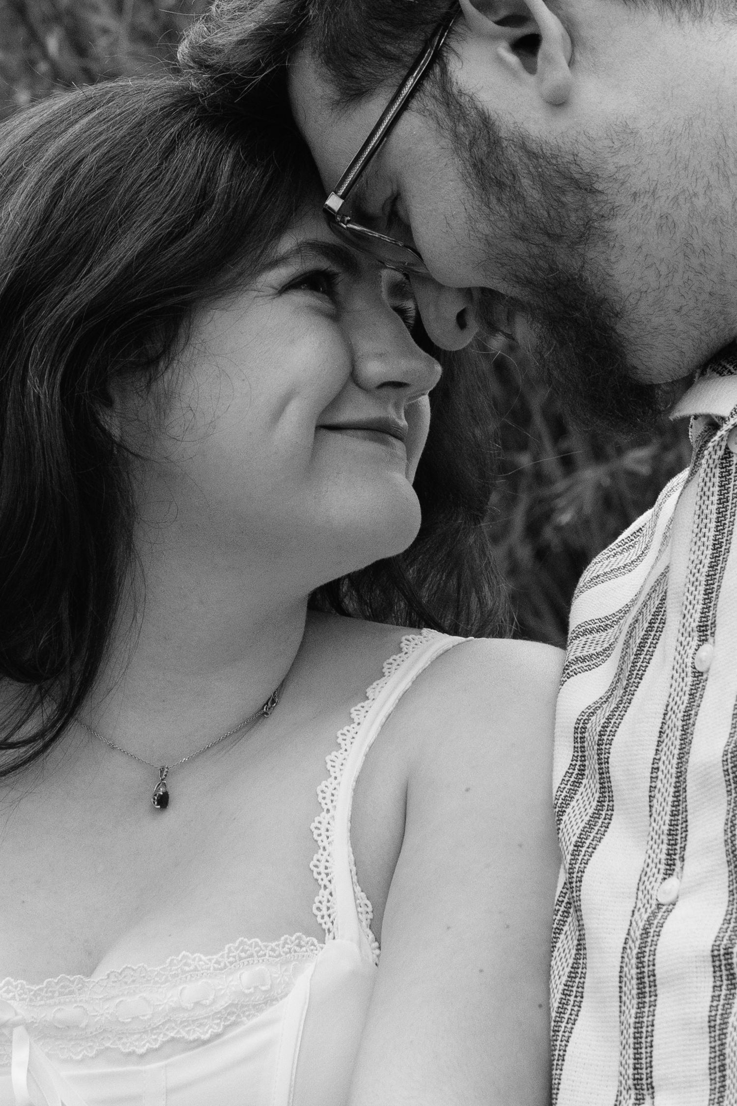 Black and white close-up of a smiling couple leaning foreheads together, woman wearing lace top and pendant necklace.