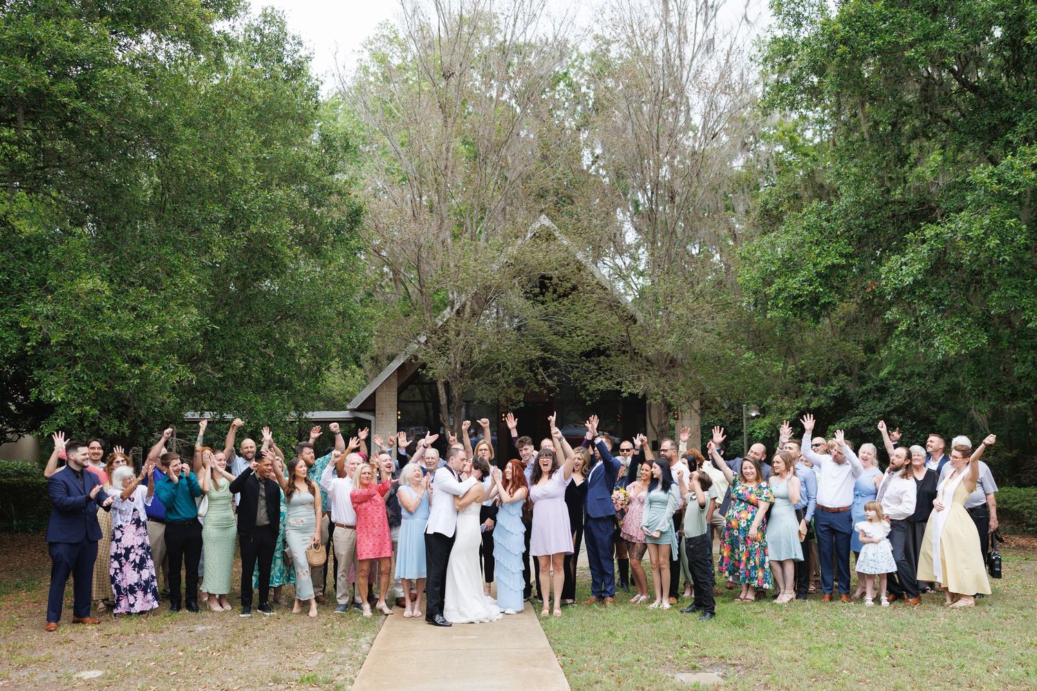 Large wedding party group photo outdoors with raised hands, celebrating in front of a rustic chapel surrounded by trees.