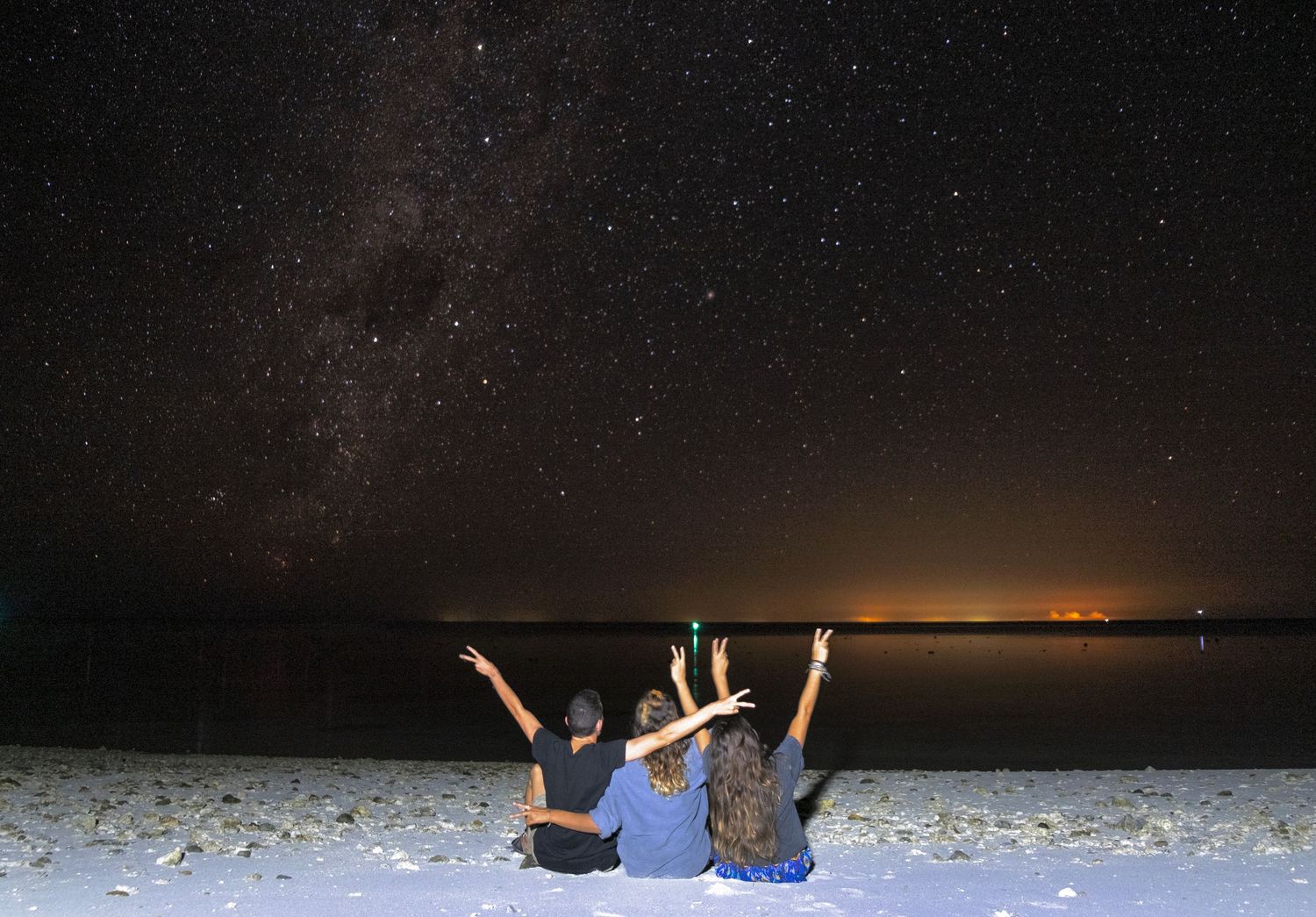 Three friends sit on a white sand beach at night, raising peace signs under a stunning Milky Way starry sky.
