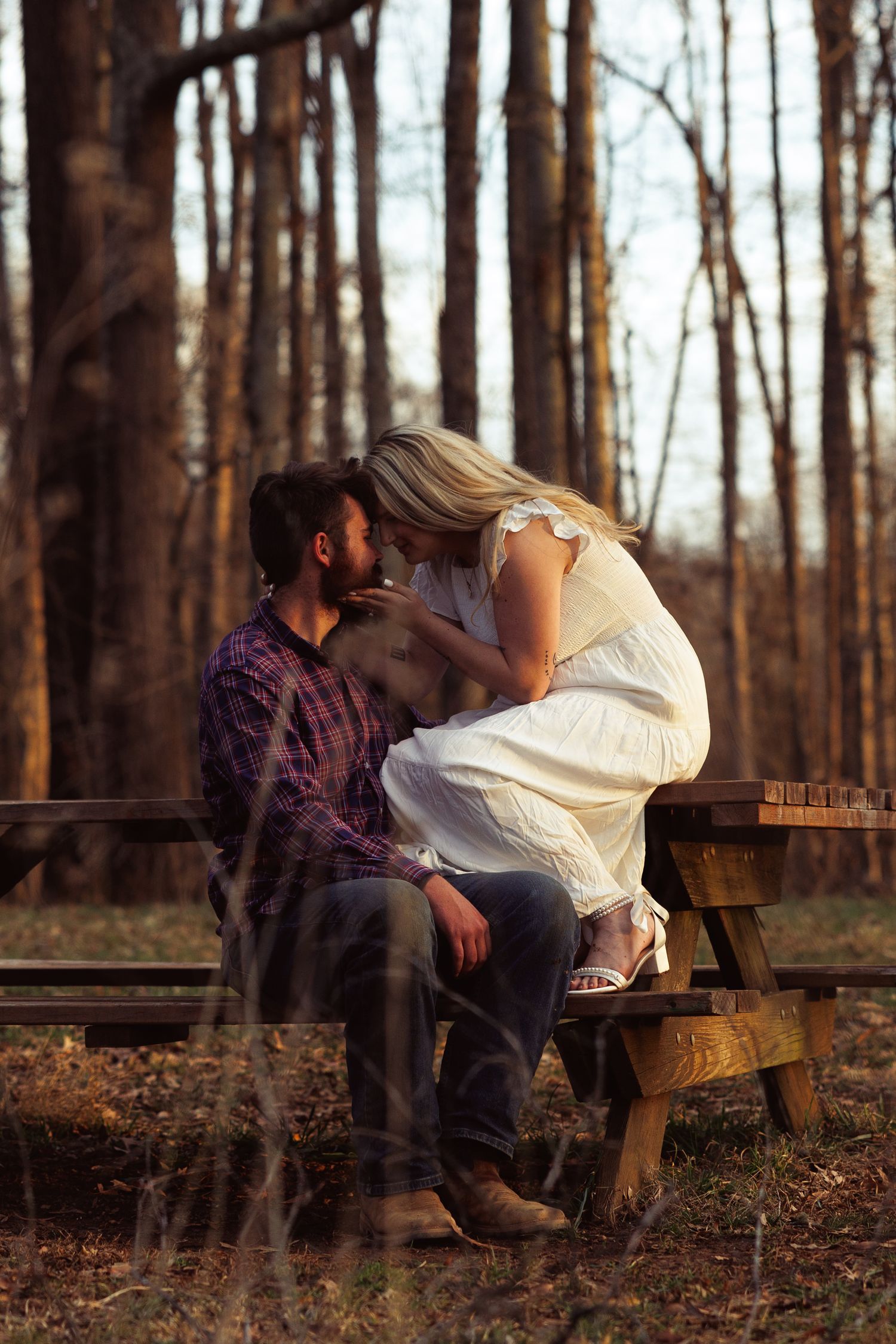 Couple kissing intimately on a rustic wooden bench in a sunlit forest setting.