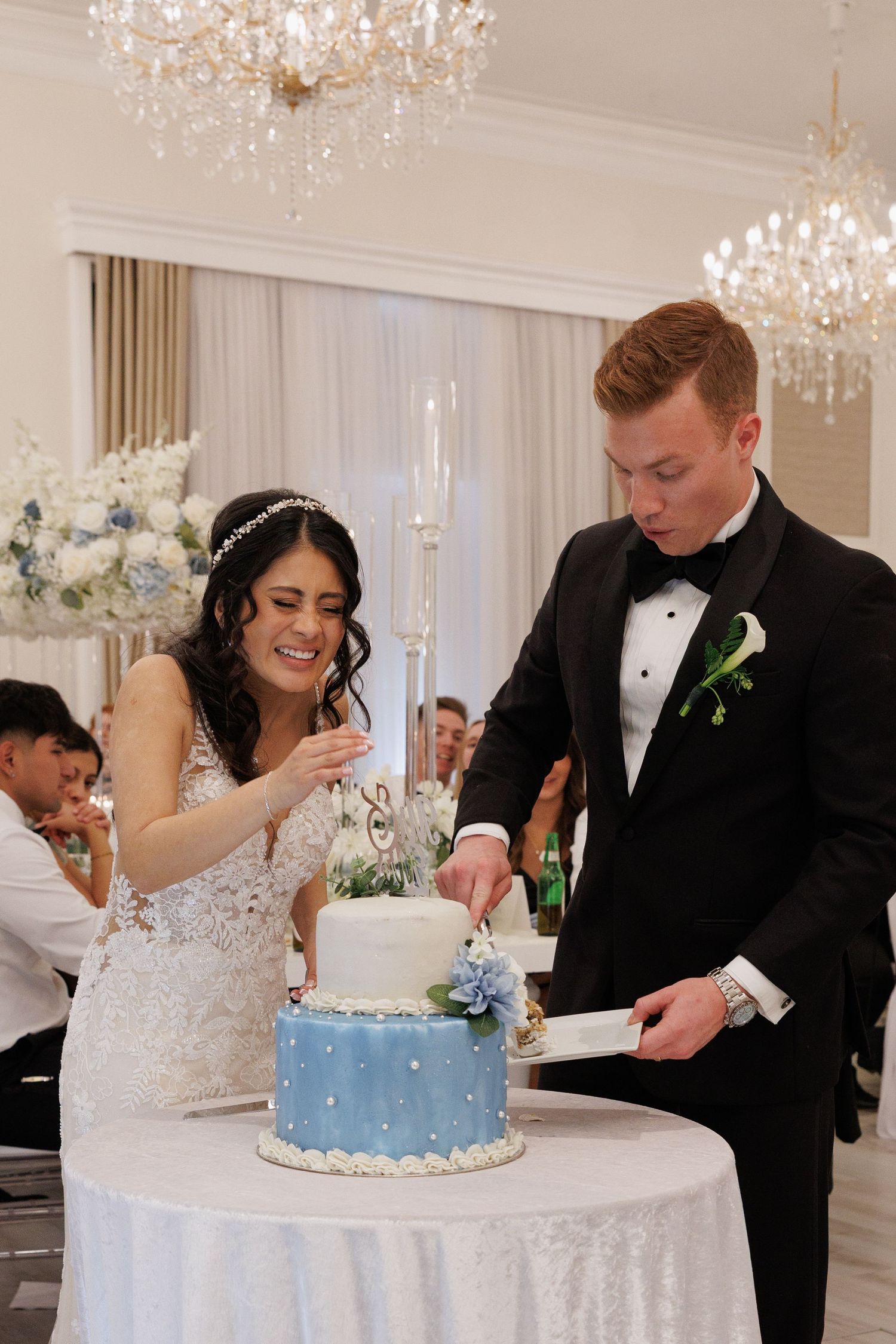 Smiling bride and groom cut a two-tier blue and white wedding cake in a chandelier-lit grand ballroom at The Yorkmont in Charlotte, NC.