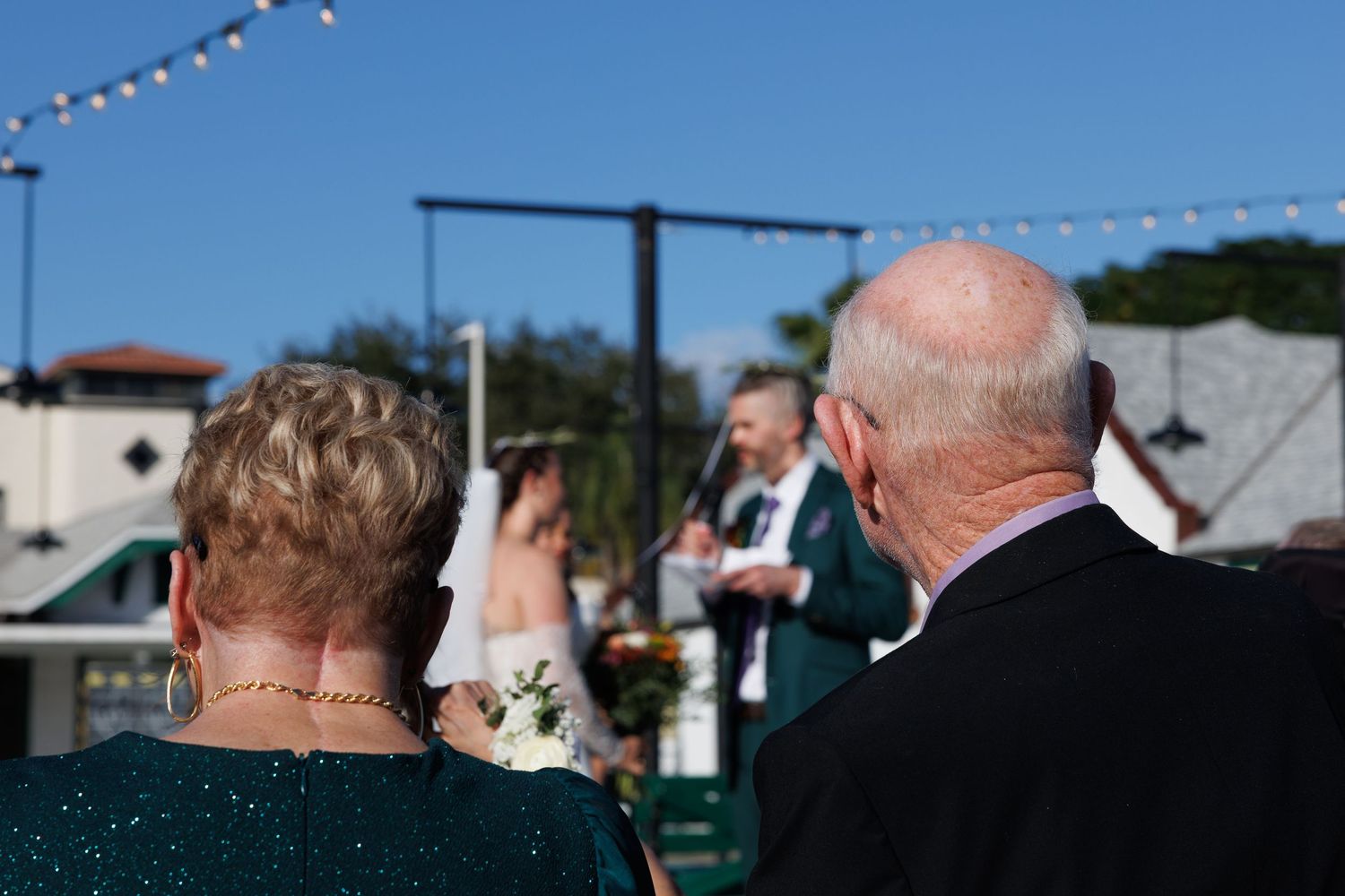 Elderly couple watching their daughter's outdoor wedding ceremony under a clear blue sky.
