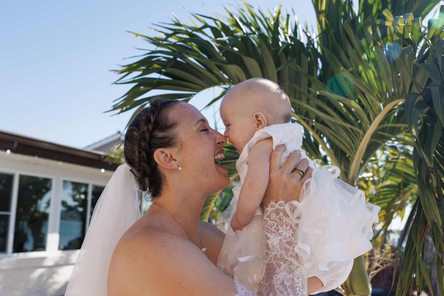 Bride in white wedding dress and veil kissing a baby outdoors near tropical palm trees on a sunny day.