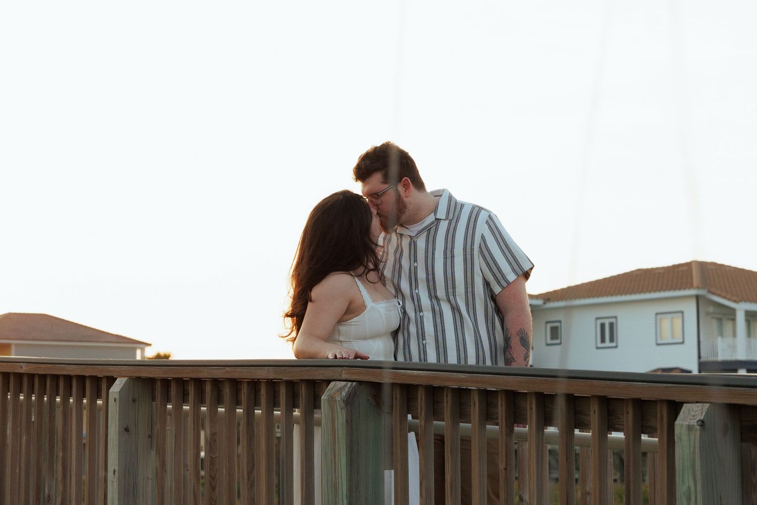 Romantic couple sharing a kiss on a wooden deck at sunset with houses in the background.