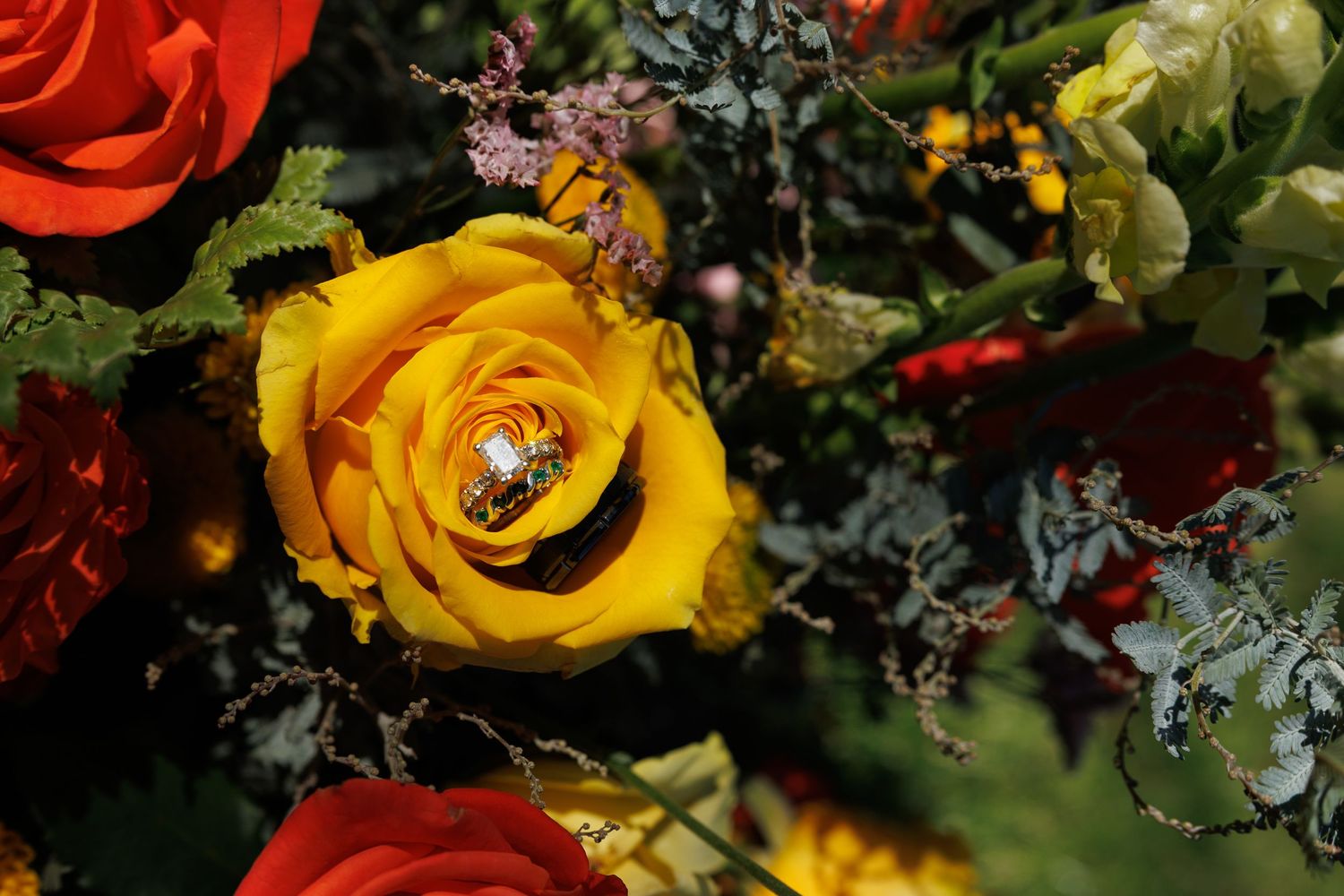 A gold engagement ring resting inside a vibrant yellow rose surrounded by red and orange roses in a wedding bouquet.