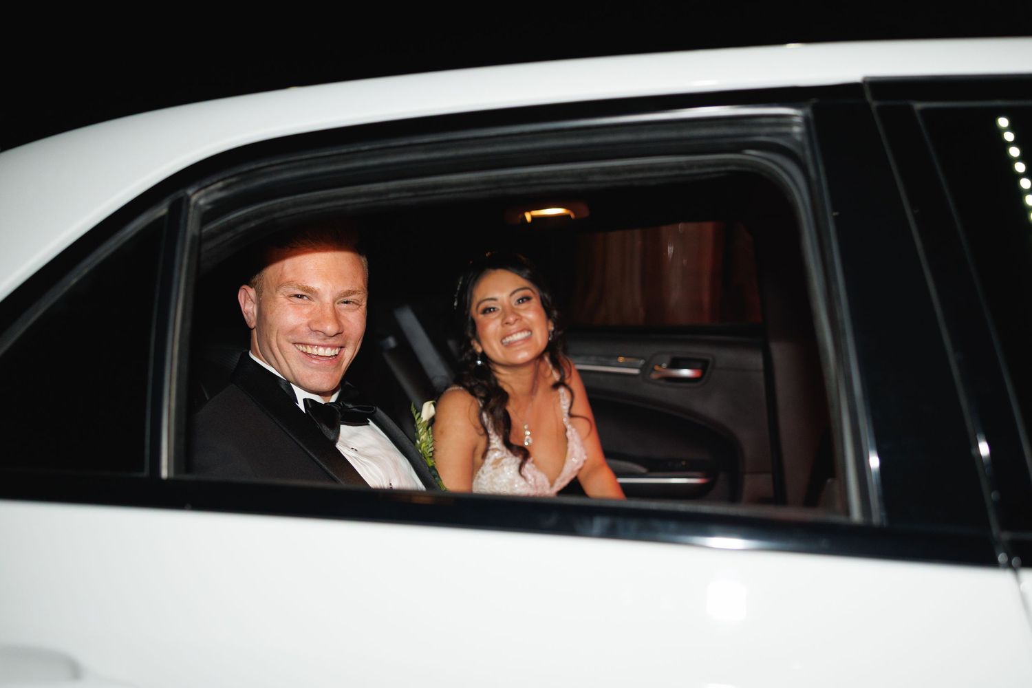 Smiling bride and groom in formal attire seated inside a white limousine on their wedding night.