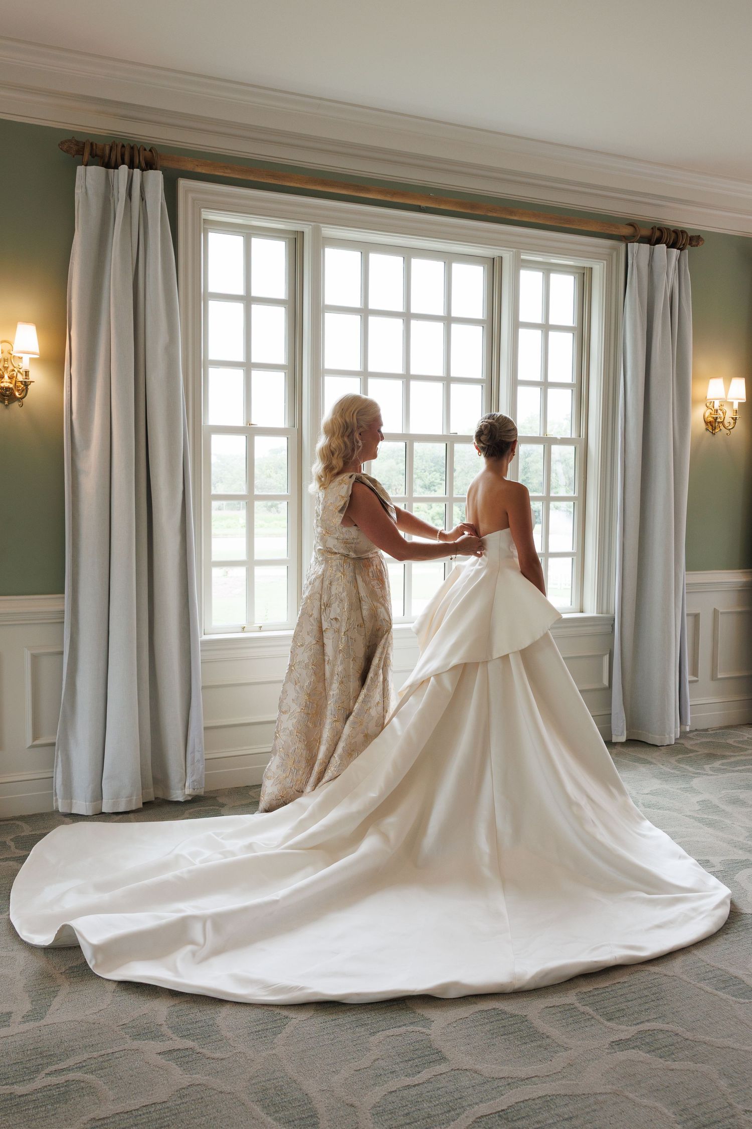 Bride and her mother share a tender moment by a large window in an elegant room before the wedding ceremony.