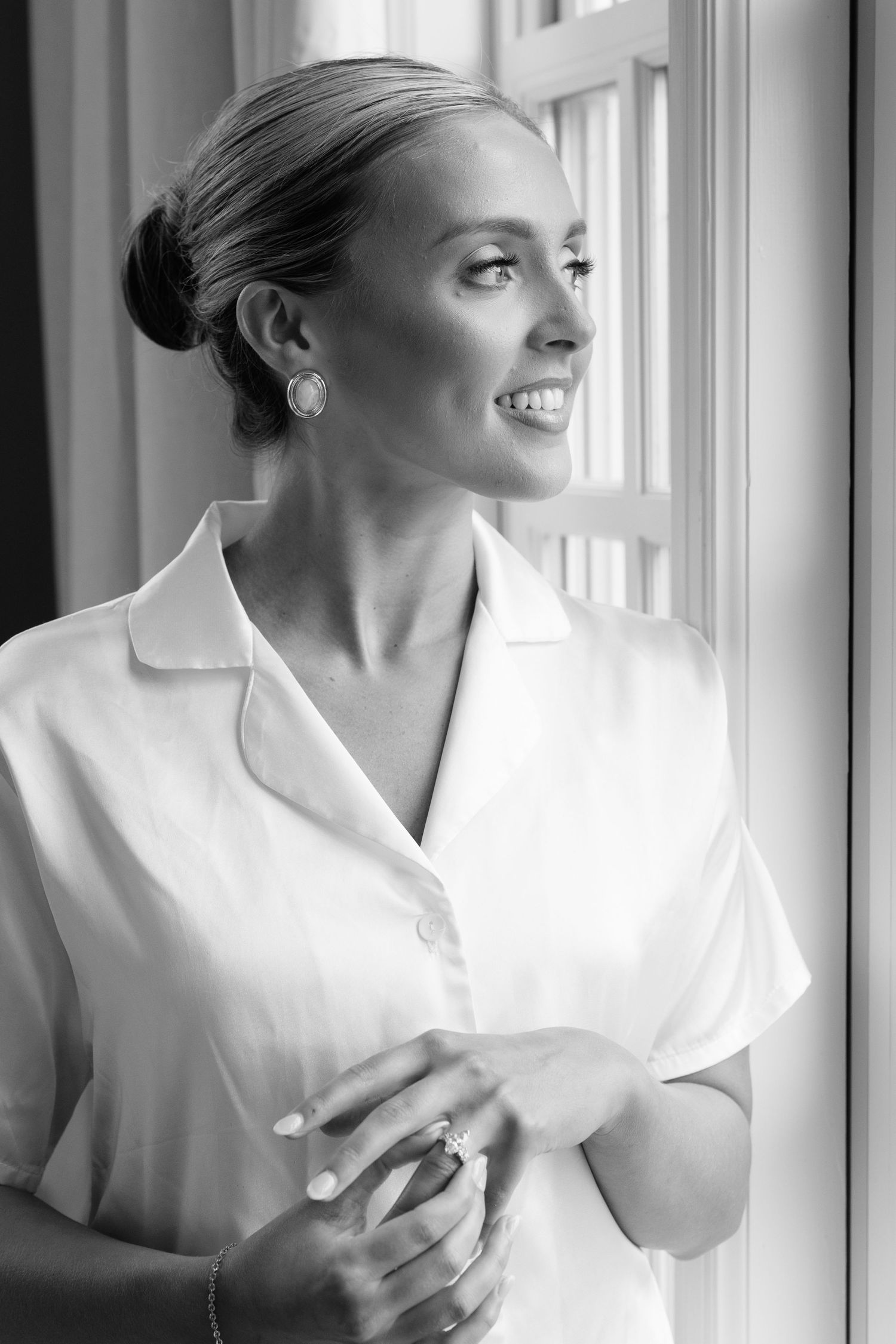 Black and white portrait of a smiling bride in white shirt gazing out a window on her wedding day.