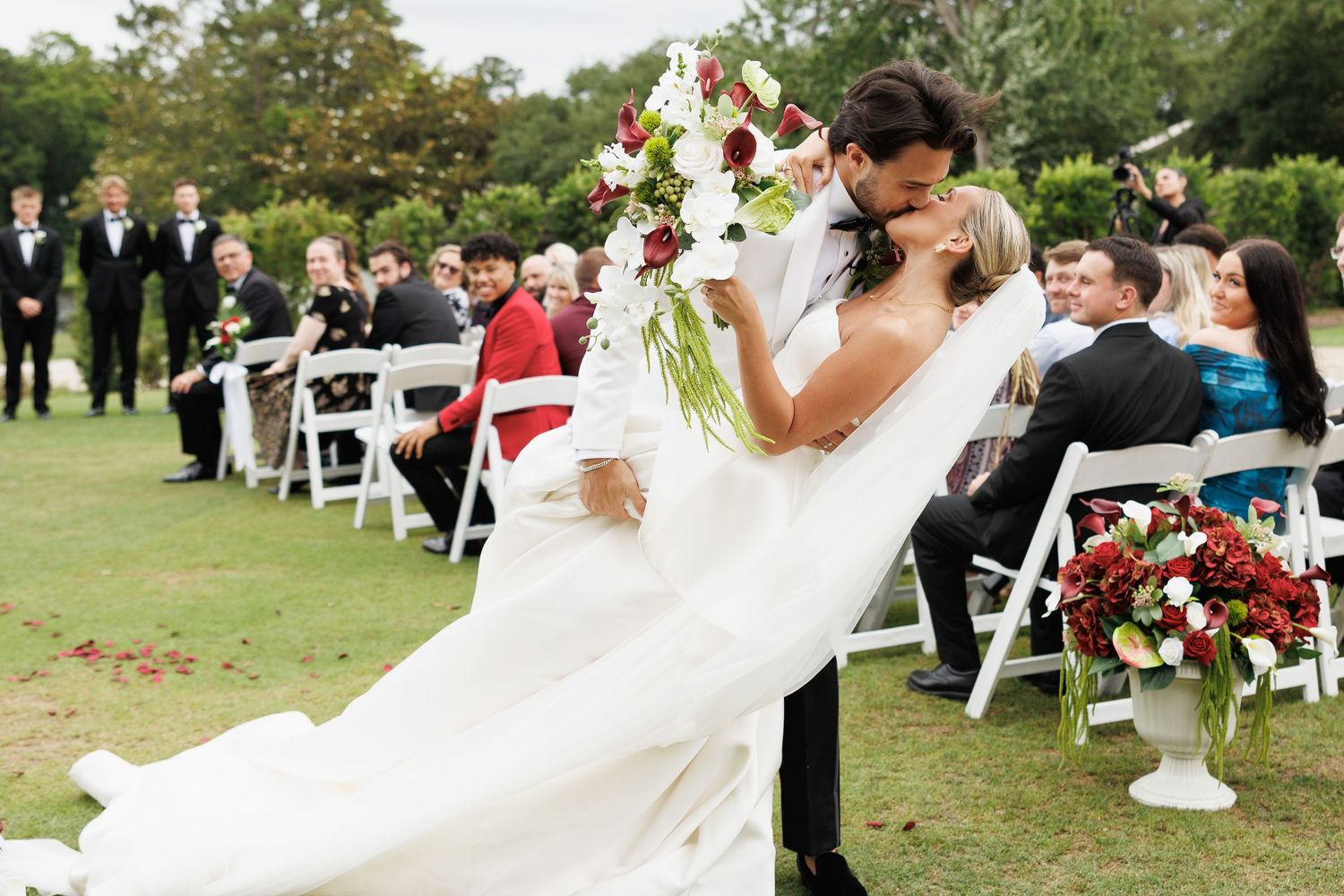 Bride and groom share a romantic dip kiss at outdoor wedding ceremony with guests seated on white chairs.