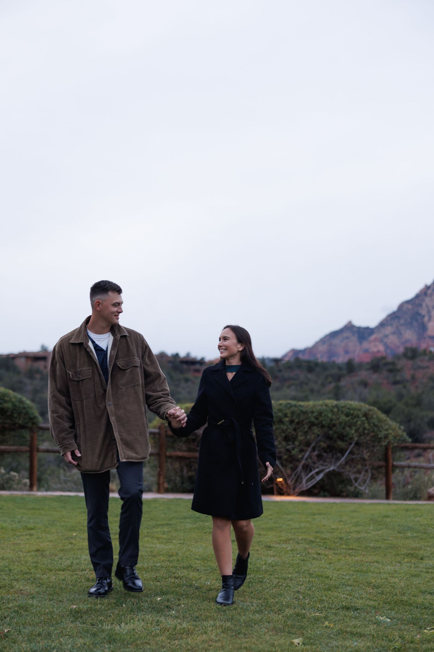 Couple holding hands and smiling at each other on green lawn with red rock mountains in background.