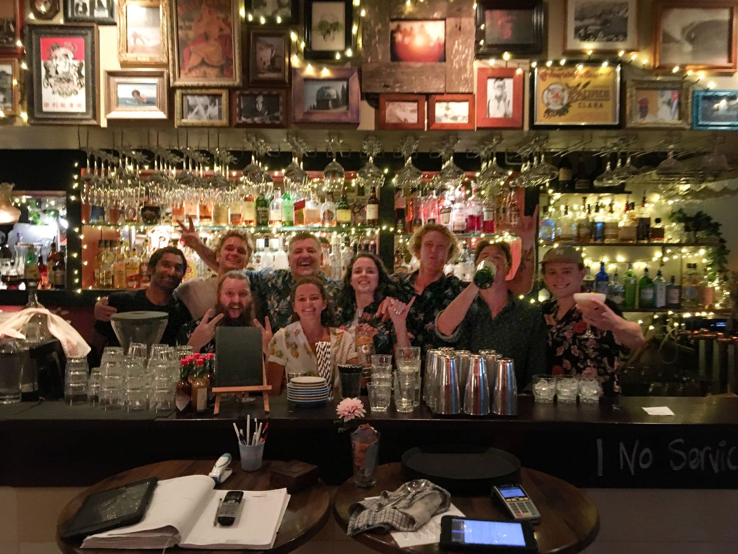 Group of smiling bartenders posing behind a decorated bar with string lights, bottles, and hanging glasses.