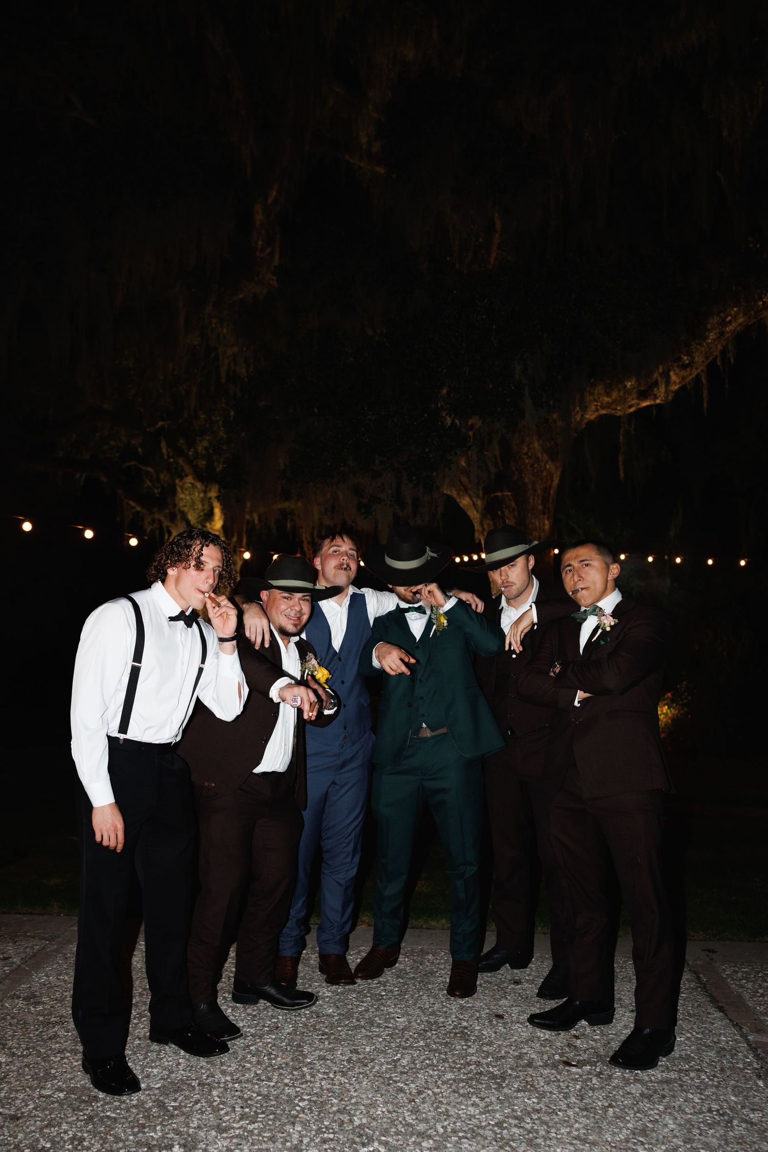 Groom celebrating with groomsmen in hats and suits under Spanish moss trees at night reception.