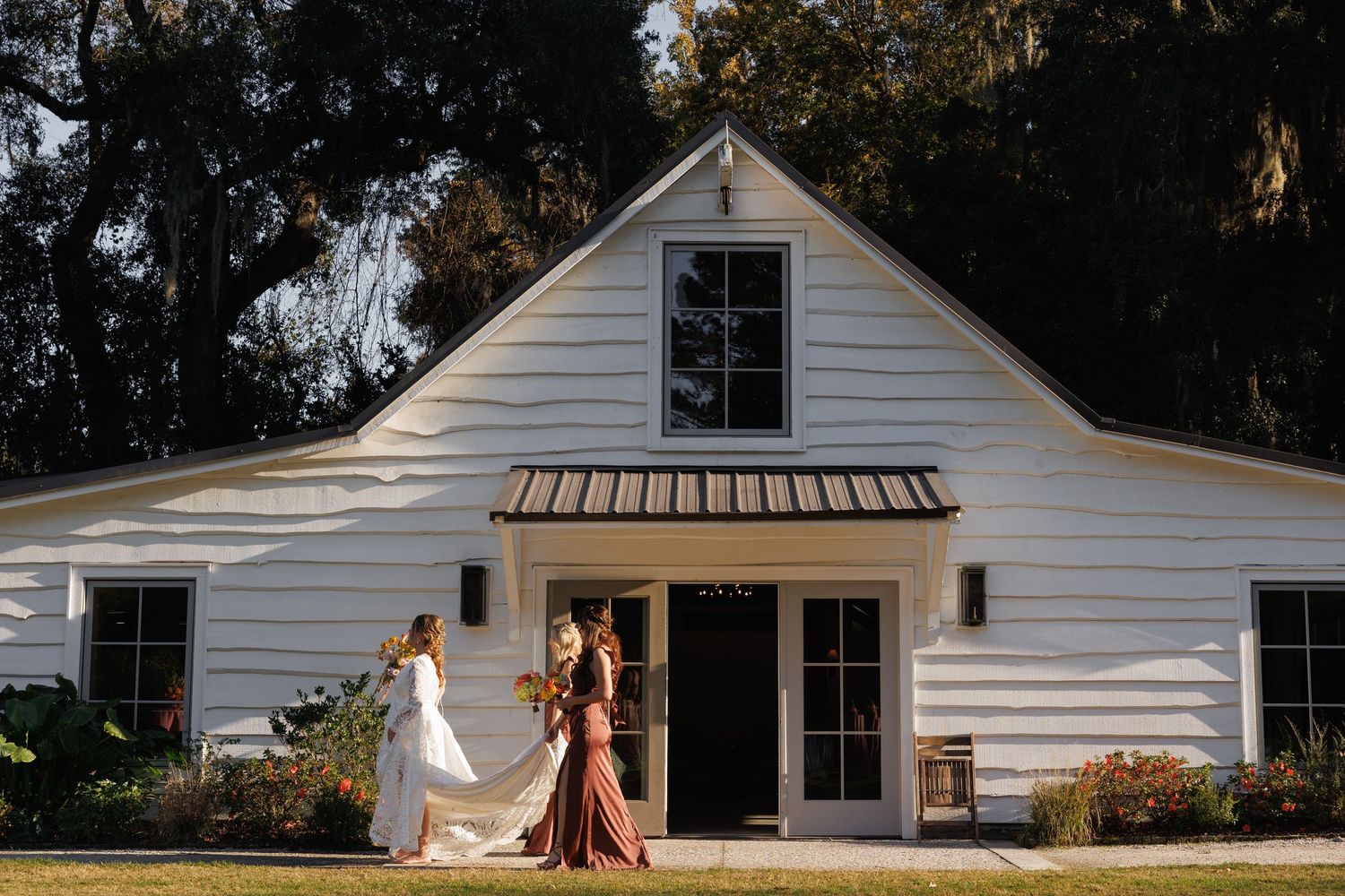 Bride and bridesmaid stand outside a white barn wedding venue surrounded by lush greenery.