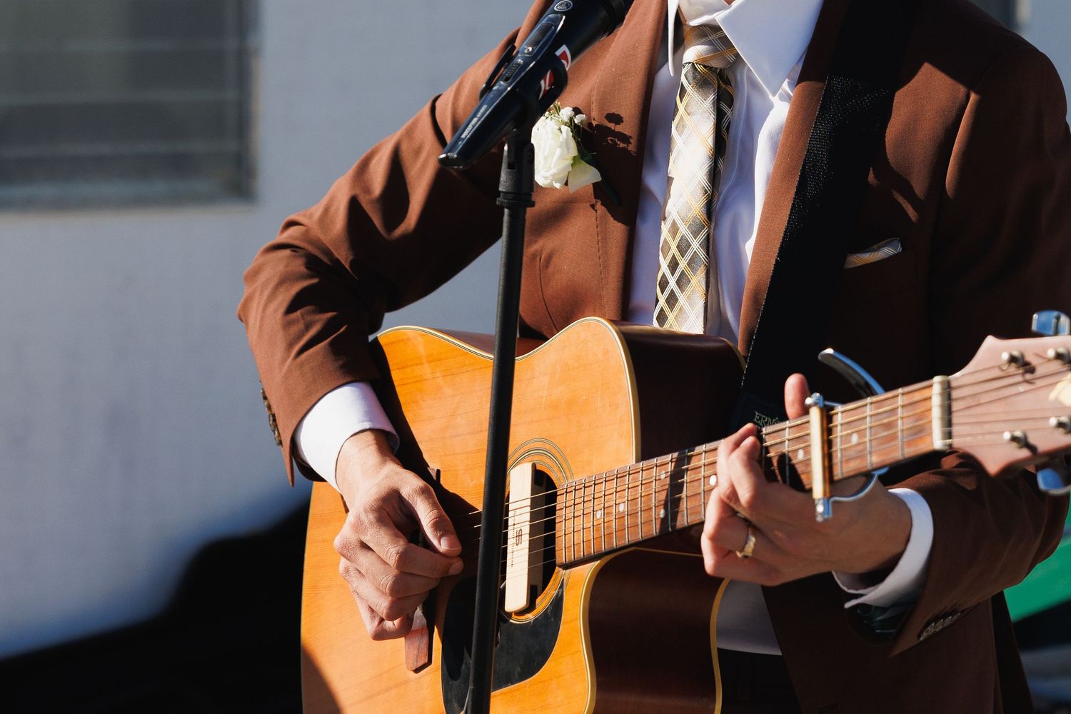 Man in brown suit playing acoustic guitar at a microphone stand, wearing a floral boutonniere.
