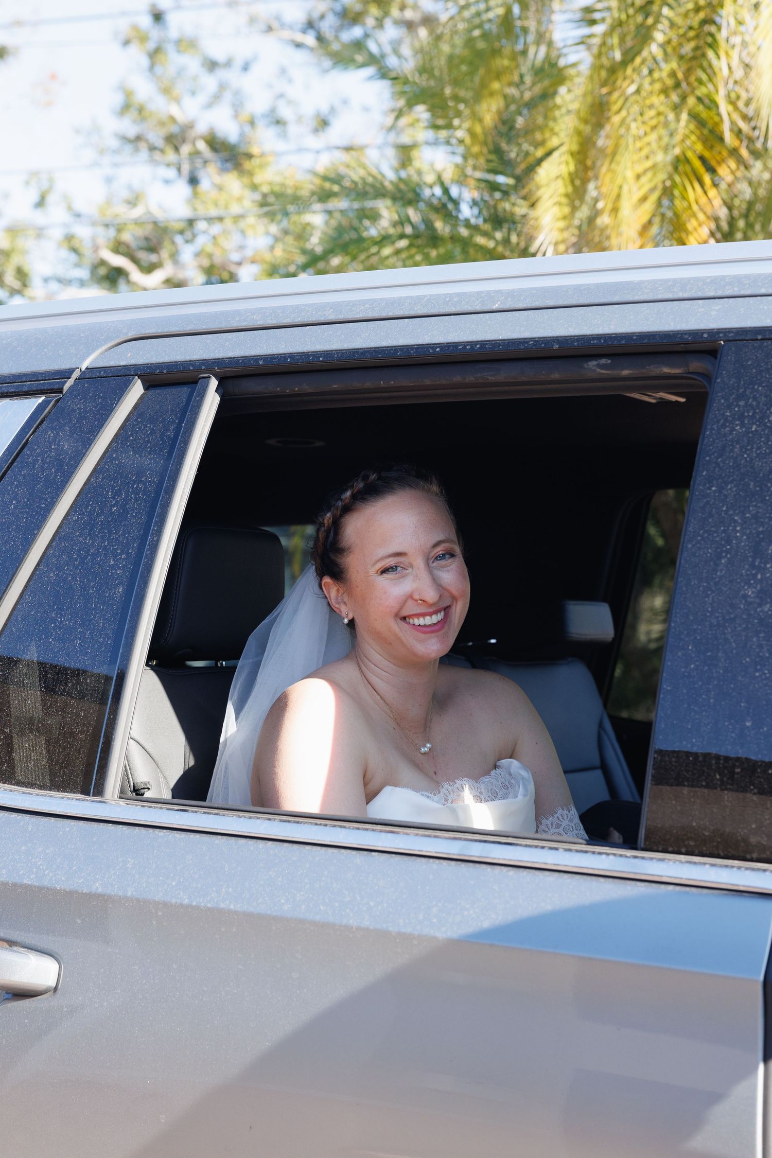 Smiling bride in white dress and veil looking out of a car window on her wedding day.