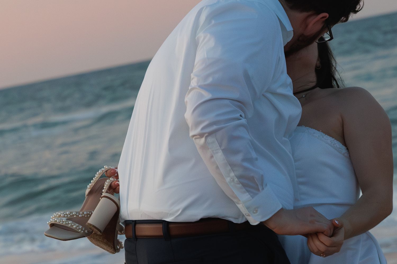 Couple embracing on a beach at sunset, groom in white shirt holding sandals, bride in strapless white dress.