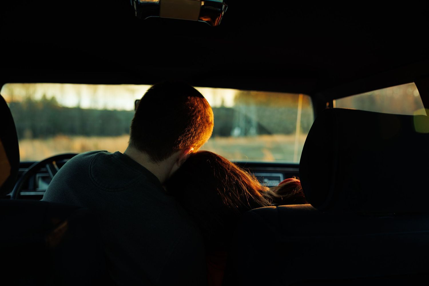 Romantic couple silhouetted from behind sharing a tender moment inside a car at sunset.